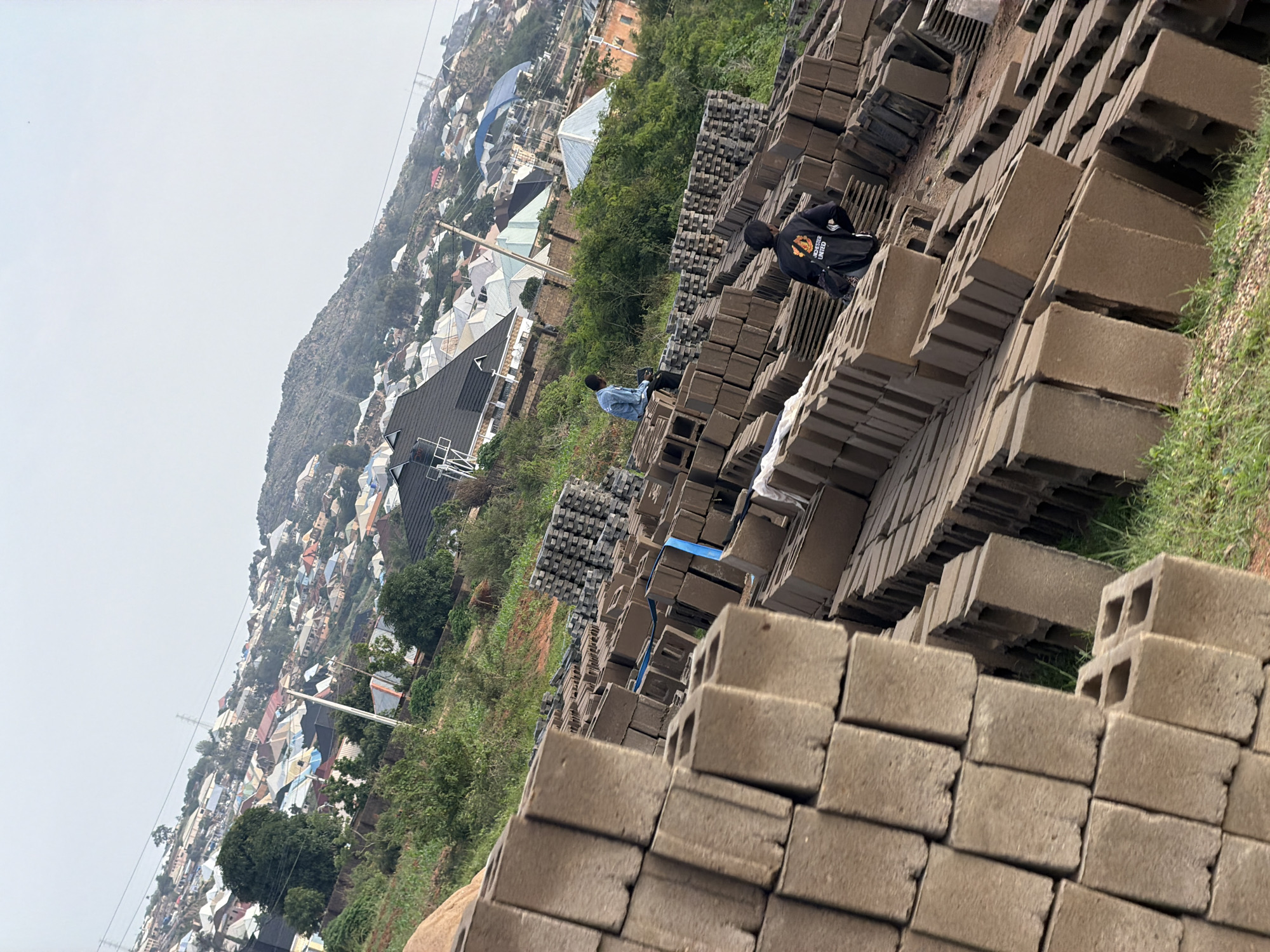 In Bukuru, Nigeria, a scene unfolds at a construction site, dominated by stacks of concrete blocks. In the foreground, these blocks are piled high, some forming neat rows and others in more haphazard arrangements. The texture of the concrete is rough and weathered. Two individuals are present: one, a man wearing a blue denim shirt, sits on a stack of blocks in the mid-ground, facing away from the camera. Another man, in a black jacket with "MANCHESTER UNITED" emblazoned on the back, stands in the mid-ground to the right, also facing away.
Behind the blocks, a verdant, grassy slope rises. Further up the slope, a sprawling residential area is visible, characterized by numerous houses with diverse roof colors, including white, blue, and red. The houses are densely packed, creating a textured urban landscape that ascends a gentle hill. The sky above is overcast and hazy, with the faint outline of power lines visible. The overall impression is one of a developing community situated against a natural, hilly backdrop.