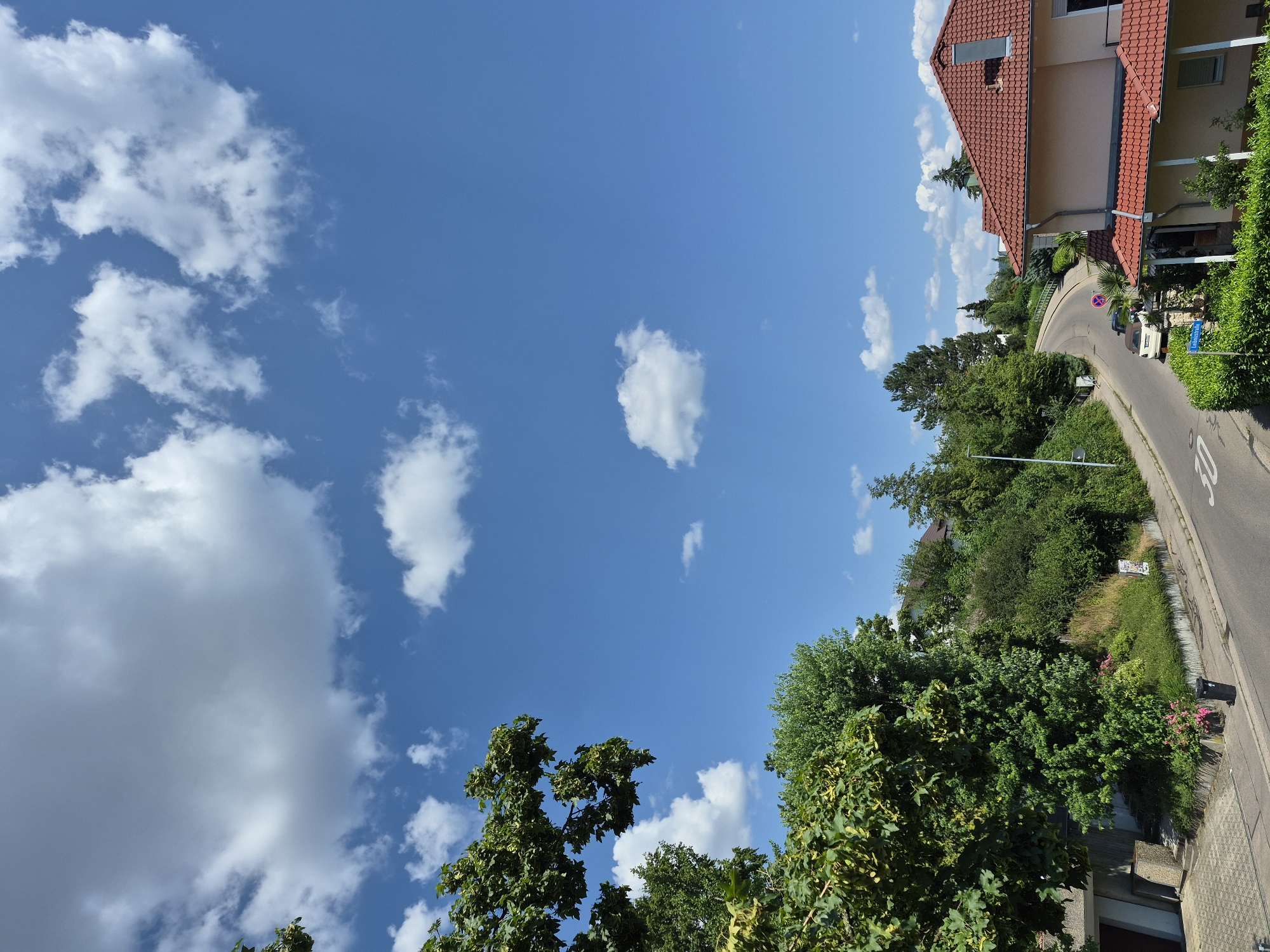 This is a vertical, outdoor, daytime photograph of a residential street scene. The sky is a vibrant blue with scattered white, fluffy clouds. Lush green trees and foliage line the right side of the frame, with a paved road curving through the scene. A house with a red tile roof is visible on the right, set back from the road. A white car is parked near the side of the road. A blue street sign reads "Lindenstraße". A speed limit sign shows "30". The overall impression is one of a peaceful, sunny day in a suburban or rural area.