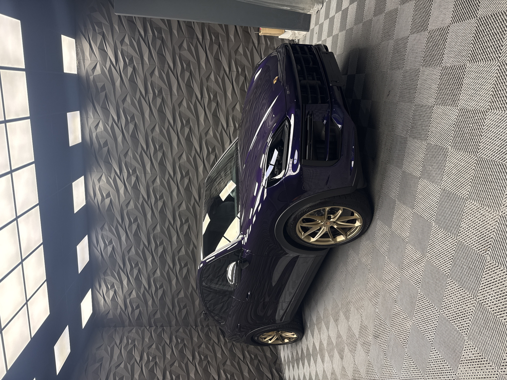 A high-gloss, dark purple Porsche SUV, with gold-colored rims, is parked indoors, angled slightly towards the right of the frame. The vehicle is positioned on a textured, light gray floor, composed of interlocking square tiles. The left side of the frame features a wall with a geometric, three-dimensional pattern in shades of gray, illuminated by a row of square lights.