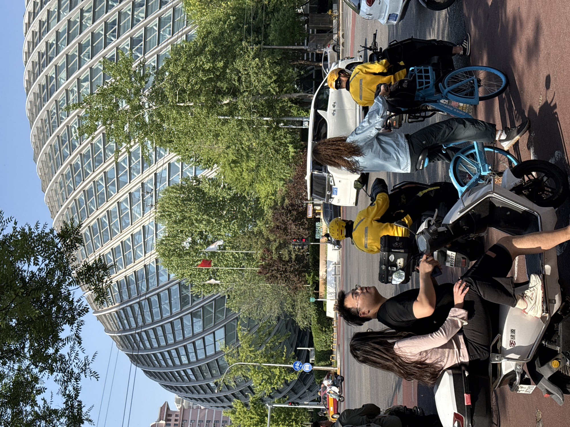 The image depicts a street scene in Chaoyang District, China. In the foreground, several people are sitting on scooters or bicycles on a road. A couple is on a scooter together, with the man wearing glasses and a black shirt, and the woman with long hair wearing a light-colored top. Two delivery drivers in yellow jackets are visible, one on a scooter and another on a blue bicycle. Cars and scooters are parked or driving on the road. In the background, there is a large, modern glass building with a distinctive curved design. Trees and flags are visible along the street. The scene is captured in daylight with clear skies.