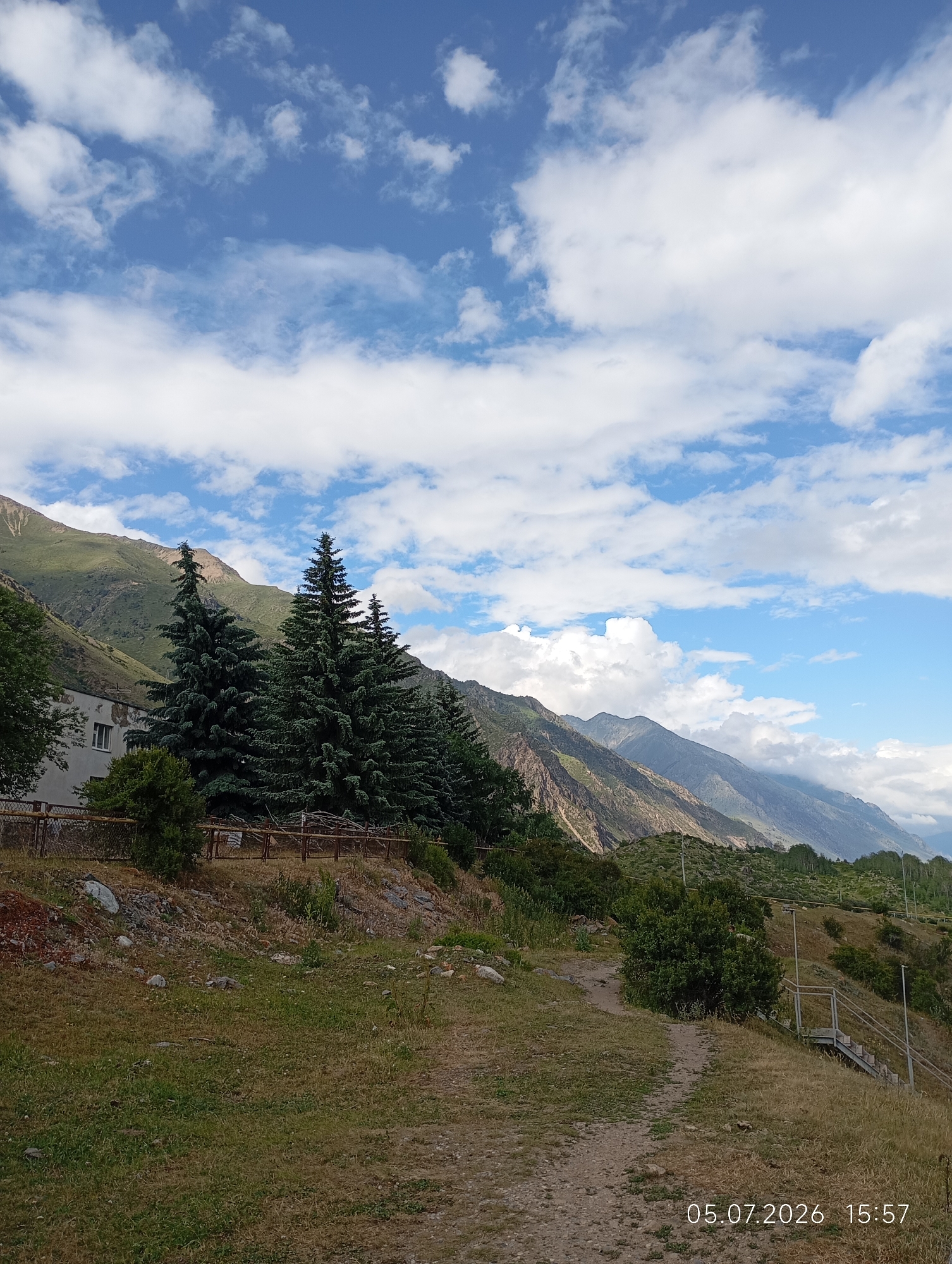 The image shows a mountainous landscape with a partly cloudy sky. In the foreground, there is a dirt path leading up a grassy slope. To the left, there is a building with a white facade and a few windows, surrounded by trees, including several tall evergreen trees. A metal fence runs along the property. In the background, rugged mountains rise, with some covered in green vegetation and others appearing rocky and barren. The overall impression is of a rural or semi-rural setting in a mountainous region. There are no people or animals visible in the image. A timestamp in the lower right corner reads "05.07.2026 15:57". This image is located in the vicinity of Neutrino, Russia.