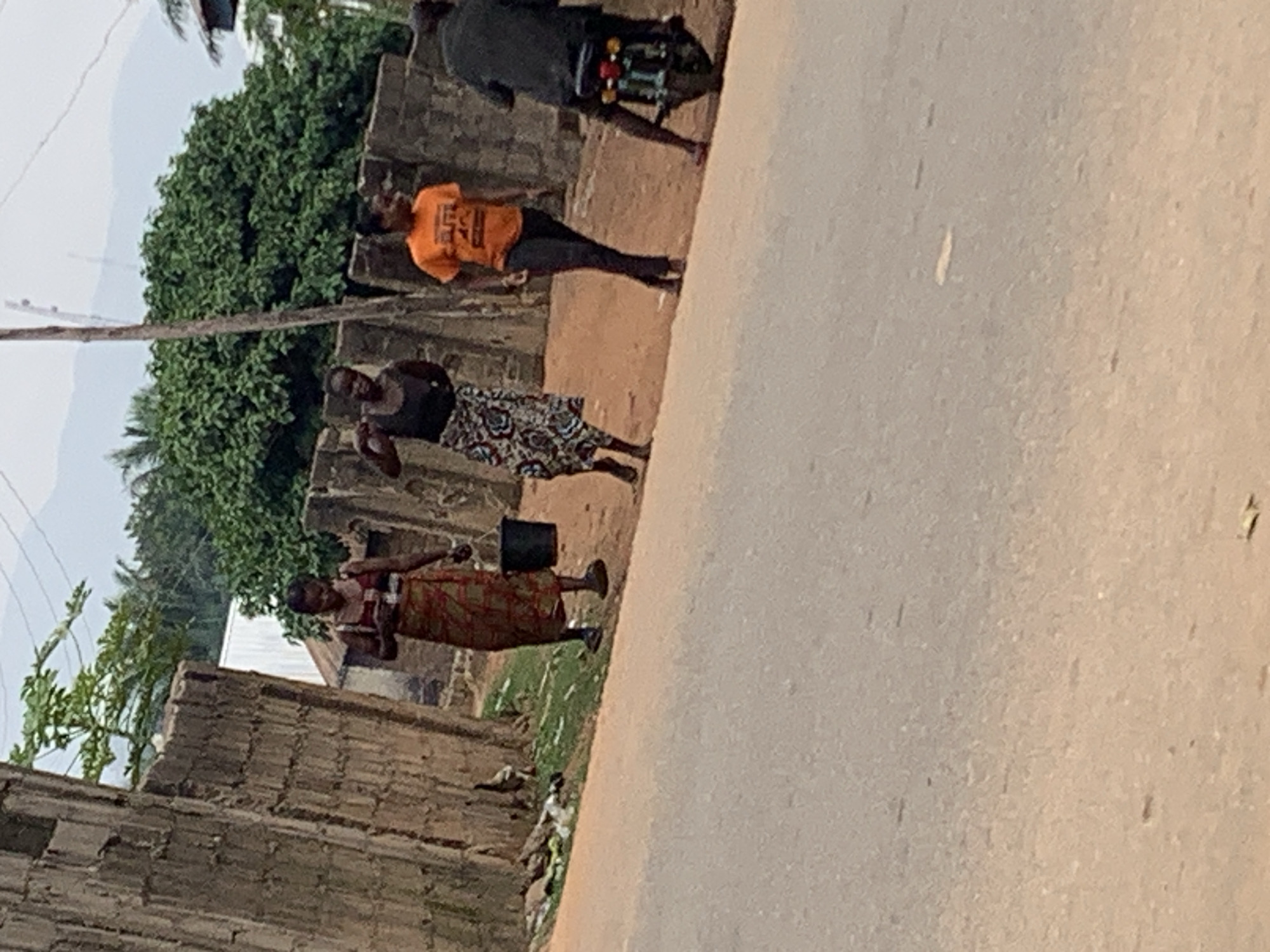 The image appears to be taken in Akwanga, Nigeria. It depicts a street scene with three people walking on the side of the road, close to a brick wall and next to some greenery. The road is wide and takes up most of the frame. A motorcycle is also visible further up the street.