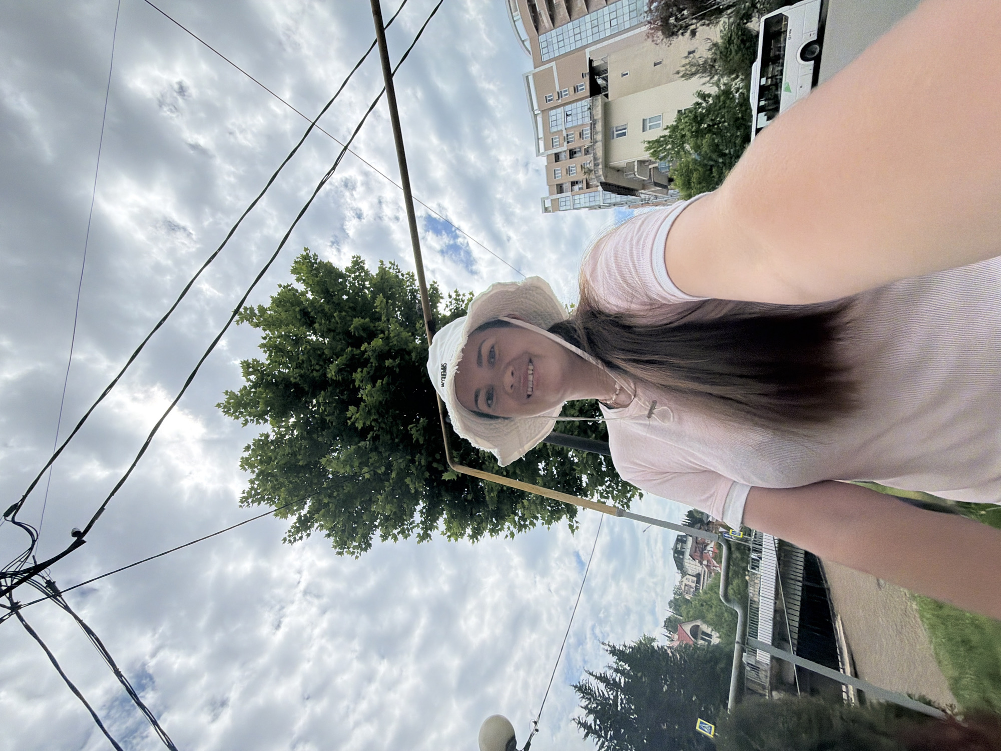 A woman is taking a selfie outdoors in Sochi, Russia, under a cloudy sky. She is wearing a white bucket hat and a light pink t-shirt. Behind her, there are several power lines crisscrossing the sky. A large green tree is visible behind her head. In the background, there are residential buildings, a street with a white bus, and some vegetation. The woman is smiling at the camera, and her arm is extended to take the photo. The overall impression is that of a casual outdoor moment in an urban environment.