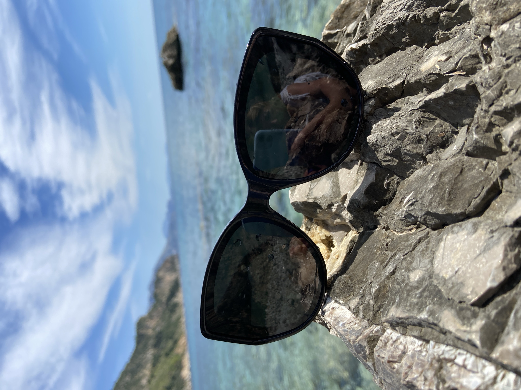 The image shows a pair of black sunglasses resting on a rock formation. The lenses reflect the surrounding landscape, including a figure of a person partially visible in the reflection, likely the photographer. The background features a coastal scene with turquoise water, a distant rocky island, and a blue sky with clouds. The rock formation is rough and textured. The location is suggested to be in or near Prijevor, Montenegro, based on the given context.