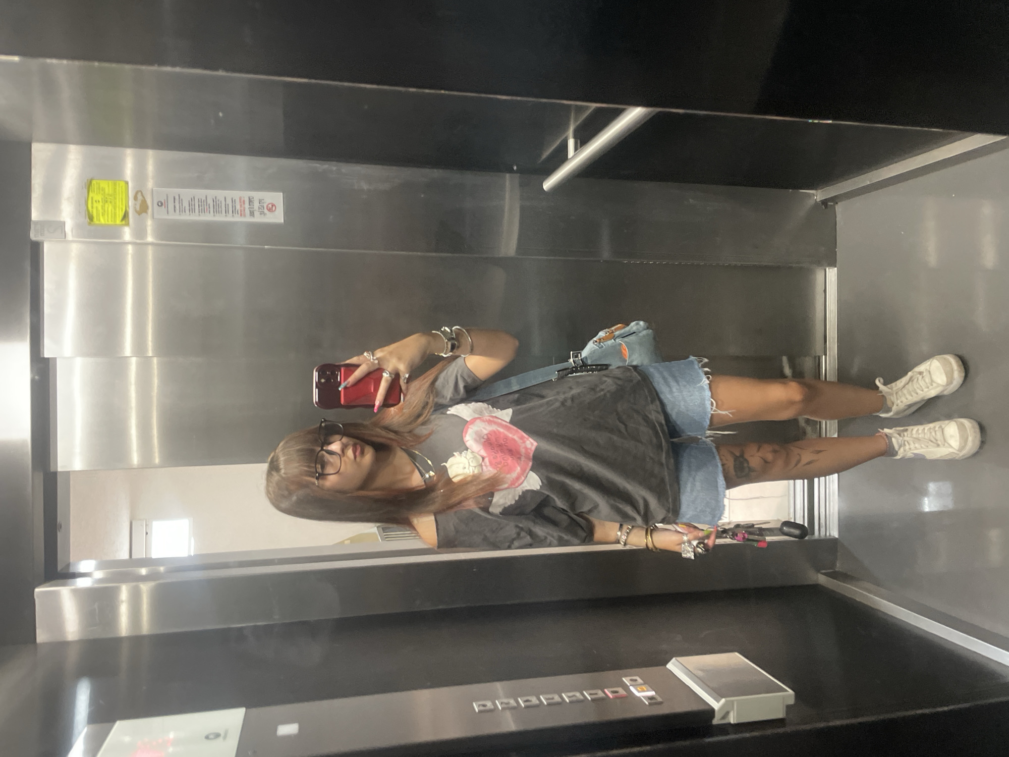 A young woman with long, brown hair and glasses is standing in an elevator, taking a mirror selfie. She is wearing an off-the-shoulder grey t-shirt with a pink heart graphic, denim shorts, and white sneakers. She has several rings on her fingers and bracelets on her wrists. A light blue crossbody bag is slung over her shoulder. Her legs have tattoos. The elevator has stainless steel walls and black paneling, with a silver handrail on the right side. There are buttons and labels on the elevator wall to the left. The floor is grey. The elevator appears to be in an urban setting, likely a building in Raanana, Israel, given the context provided.