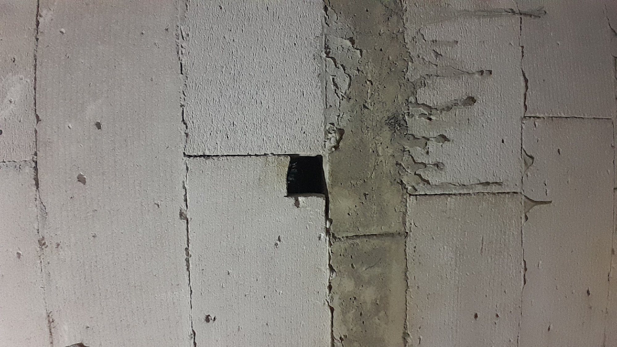 The image shows a close-up of a wall constructed from light-colored, rectangular blocks, possibly concrete or aerated concrete. There is a dark, square-shaped opening in the wall, suggesting a hole or vent. The blocks are arranged in horizontal rows with visible mortar lines. Below the opening, there is a band of rough, darker material, possibly cement or concrete, with some dripping or irregular textures. The overall scene appears to be an unfinished construction or an outdoor wall. The location is identified as Sumberarum, Indonesia.
