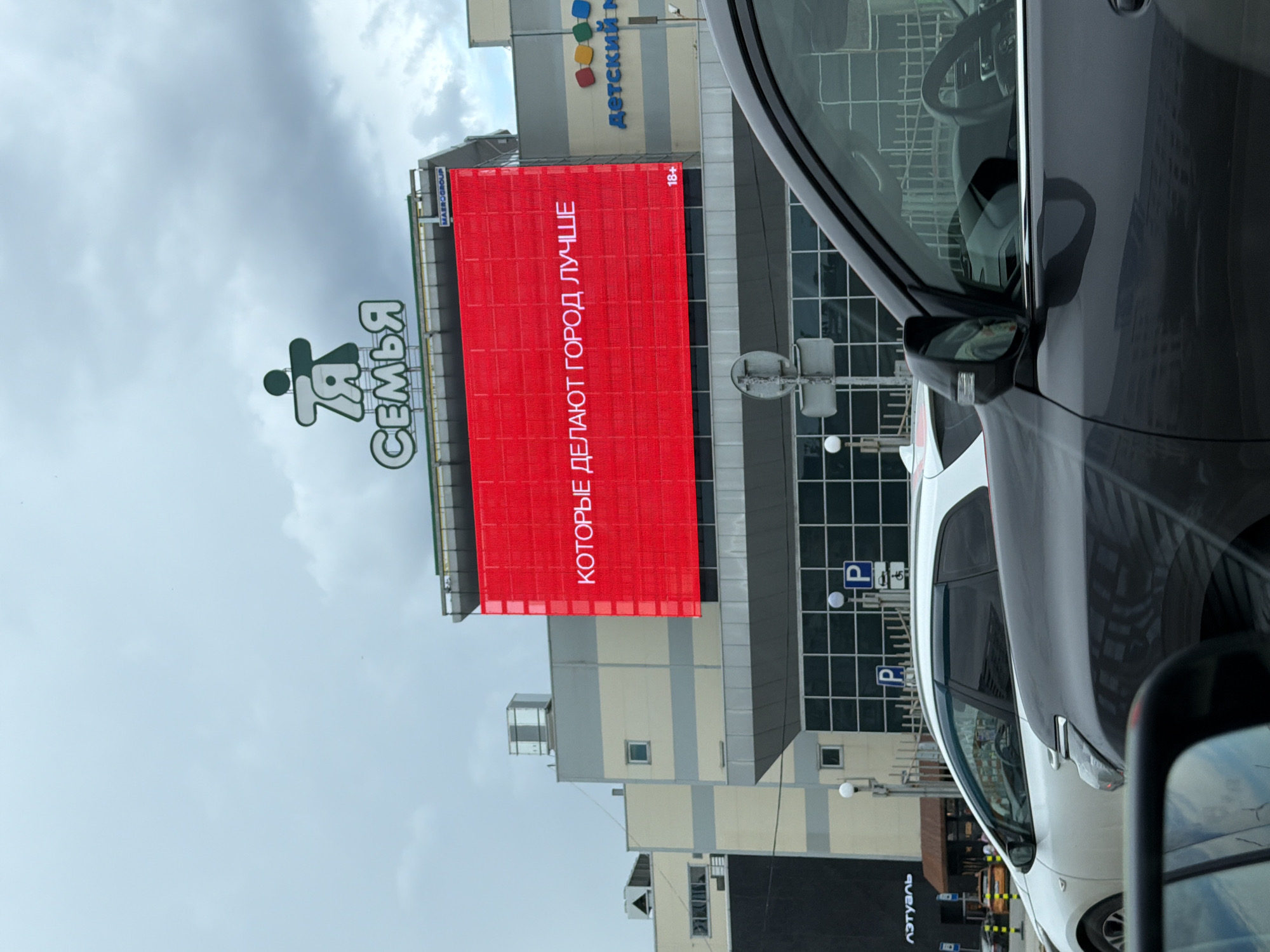 The media file captures a scene in Perm, Russia, featuring a large red billboard on the side of a modern building. The billboard displays text in white letters, oriented vertically and appearing to be in Russian. The text reads "КОТОРЫЕ ДЕЛАЮТ ГОРОД ЛУЧШЕ" which translates to "THAT MAKE THE CITY BETTER." To the left of the billboard, a logo featuring stylized figures and the word "СЕМЬЯ" (Family) is visible. The building itself has a contemporary architectural style with multiple windows and sections.
In the foreground, the side of a dark-colored car is prominent, with its interior, including a steering wheel and dashboard, partially visible through the window. Next to the dark car, the rear of a white car is also in view. The scene appears to be a street or parking area in front of the building.
Above the building, a cloudy sky suggests overcast weather. The lighting indicates it could be daytime, but the lack of direct sunlight due to the clouds makes it difficult to pinpoint the exact time. The overall atmosphere is urban and functional, with commercial signage and vehicles suggesting a public or commercial space. Visible signs include parking symbols ("P") on the building's facade. A sign with "ДЕТСКИЙ М" and colorful square shapes, likely indicating a children's store, is also present on the building. Another sign near the top of the billboard reads "18+", indicating age restriction. The presence of text in Russian confirms the location in Russia.