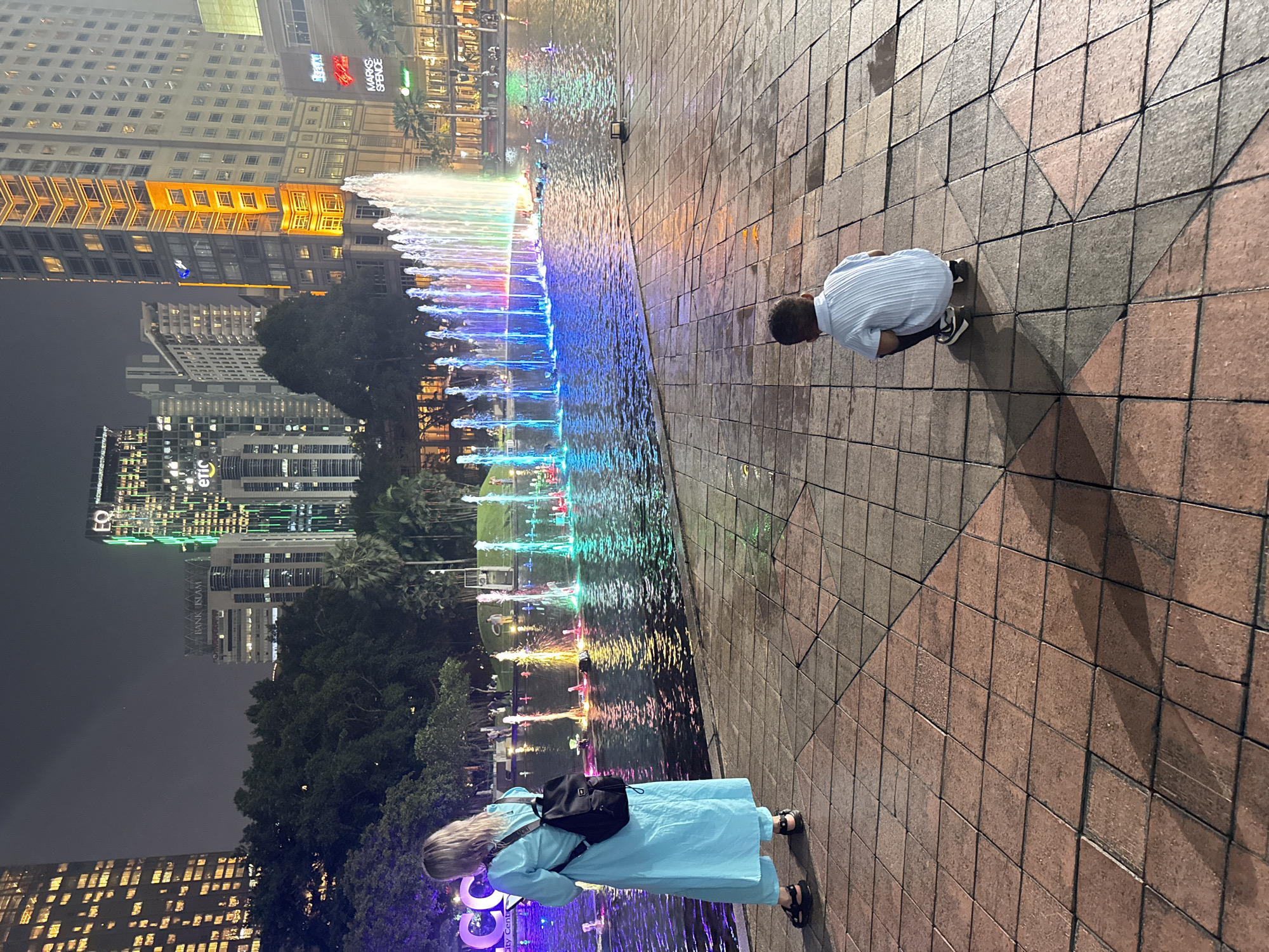 An outdoor night scene in Kuala Lumpur, Malaysia, features two individuals observing a vibrant, illuminated fountain display in a city park setting. The sky is dark, indicating evening.
In the foreground, a person with short dark hair, wearing a light blue striped short-sleeved shirt and dark sneakers, crouches on a tiled walkway. They are positioned with their back partially to the camera, looking down towards the colorful fountain. Further left, with their back completely to the viewer, stands another individual with blonde hair, wearing a long, loose-fitting light blue dress and black sandals, carrying a black backpack. This person is also looking towards the fountain.
The large fountain occupies the midground, with numerous jets of water rising and falling, illuminated in a dynamic array of colors including blue, green, yellow, pink, and purple. The lights reflect on the water's surface and the surrounding tiled ground.
In the background, a modern city skyline rises, dominated by tall buildings with many windows, some of which are brightly lit. Visible text on building signs includes "MARKS & SPENCER" and "BANK ISLAM." Other partially visible text includes "EQ" and "eric" on a building further in the distance. Lush trees and foliage are visible between the fountain area and the city buildings. The ground where the people stand is paved with square and triangular tiles in shades of brown and grey. The overall atmosphere appears leisurely, with people enjoying the city's nighttime attractions.
