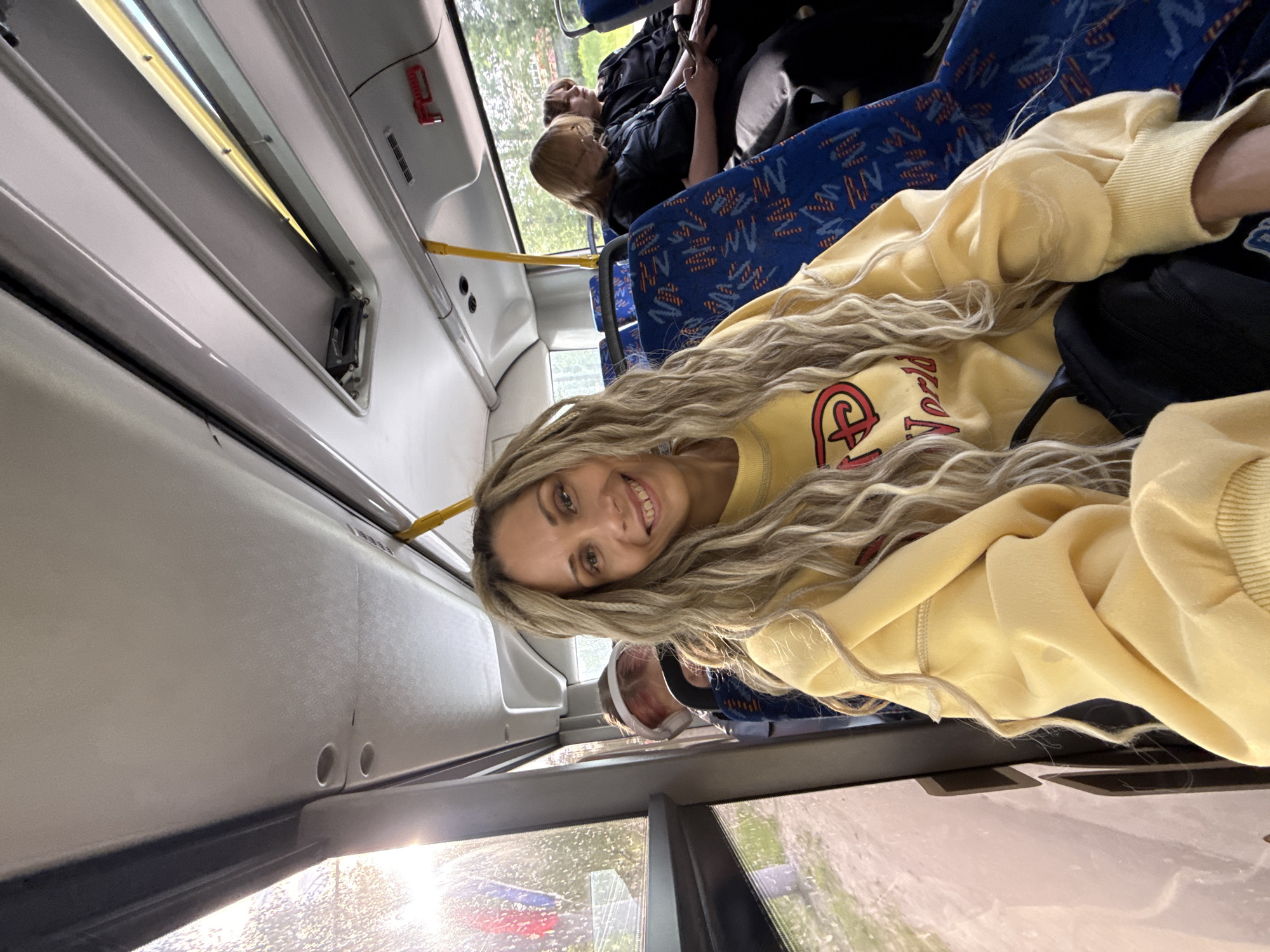 A woman with long, wavy blonde hair and a yellow sweatshirt is on a bus, likely in transit. The bus interior is visible, featuring blue patterned seats and handrails. Other passengers are present. Through the bus window, trees and a street scene are visible. The location, according to the prompt, is Rusanovo, Russia, though this cannot be independently verified from the image. The time of day appears to be daytime, with natural light illuminating the scene. The weather outside is not explicitly clear, but there is a hint of moisture or rain on the window.