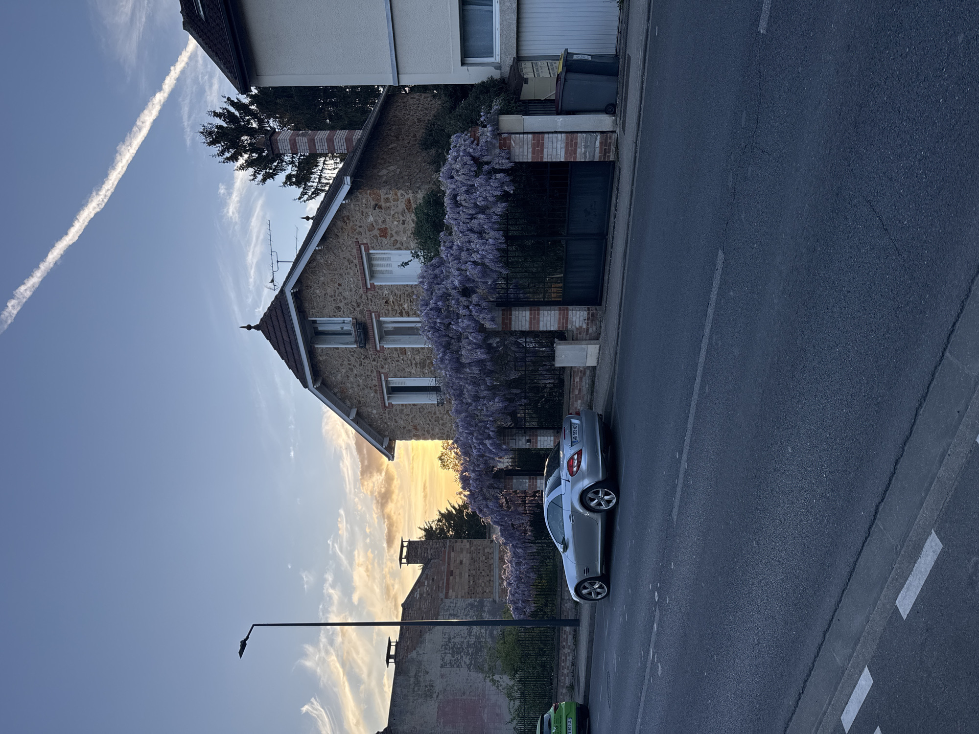 The image depicts a serene street scene in Bures-sur-Yvette, France, observed during late afternoon or early evening, characterized by a mix of natural light and twilight hues. The sky is predominantly clear blue, adorned with wispy white clouds, particularly warm and golden towards the horizon on the right, suggesting sunset. A prominent white contrail from an aircraft diagonally streaks across the upper left portion of the sky.
On the left, a charming stone house with a pitched, tiled roof stands out. It features multiple windows, some fitted with closed white shutters, and a brick chimney. A luxuriant purple wisteria vine heavily drapes over the fence and part of the house, adding a striking splash of color to the architectural elements. Behind this house, a tall conifer tree is visible. Adjacent to it, a building with a white facade and a garage door is present, accompanied by two waste bins. Further along the street, an older, partially deteriorated building with exposed brickwork can be seen.
An asphalt road runs across the foreground, marked with clear white lines. A silver two-door car, a Mercedes-Benz SLK-Class, is parked on the street. Its French license plate, "DB-983-GW," is distinctly visible, including the "F" country code. A small part of a green car is also visible further down the street on the left. A black streetlamp stands in the foreground, and an antenna is mounted on the roof of the stone house. The scene conveys a peaceful, quiet atmosphere in a residential setting.