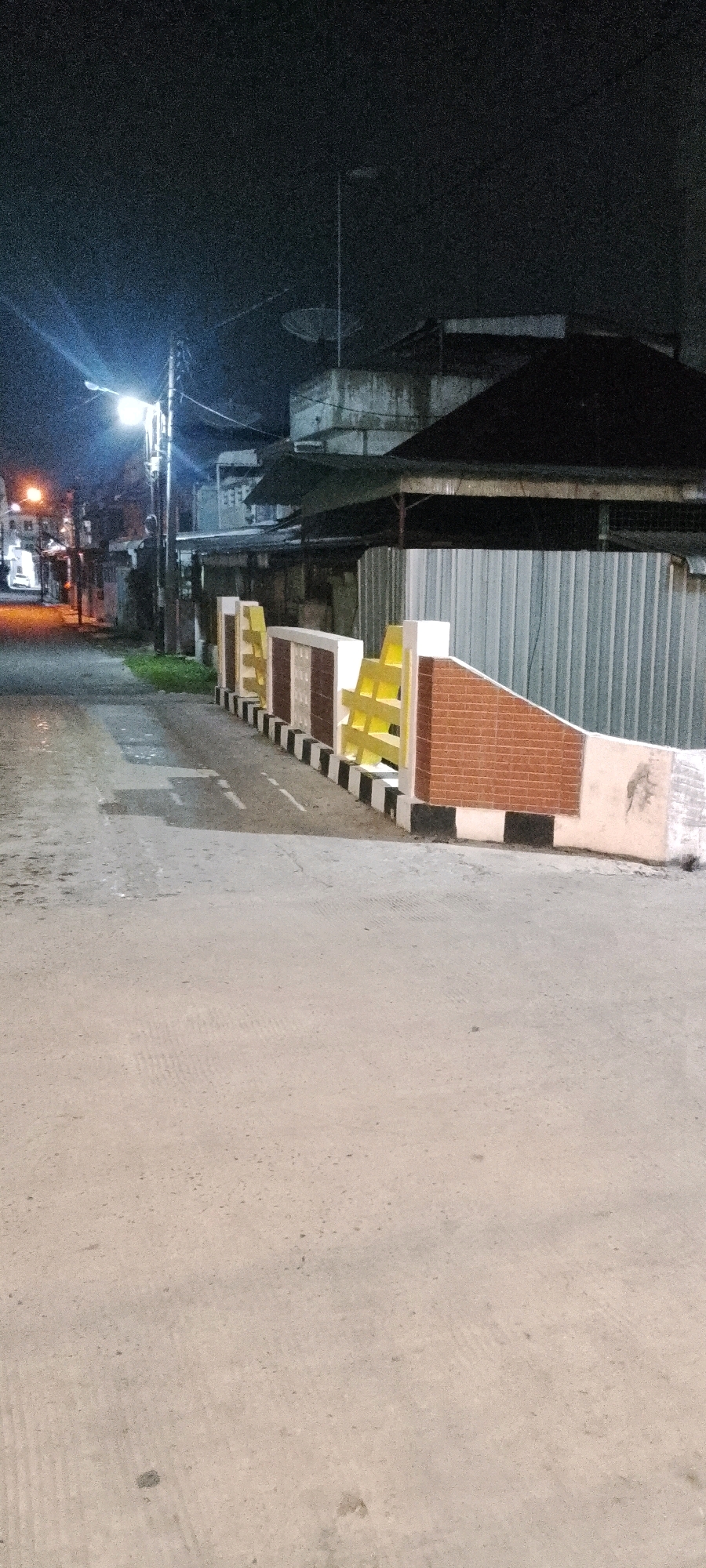 The image shows a street scene at night in the City of Medan, Indonesia. The street is paved and appears to be made of asphalt or concrete, with some visible markings. A low decorative wall with yellow and brick-patterned sections runs along the right side of the street. Behind the wall, a corrugated metal fence is visible, and beyond that, several buildings with dark roofs and some with exposed concrete sections. A satellite dish is mounted on the roof of one of the buildings. A streetlamp illuminates the scene from the left side of the image, casting light onto the street and the adjacent buildings. The overall atmosphere is dark and quiet, typical of a street at night. There are no people or vehicles visible in the frame.