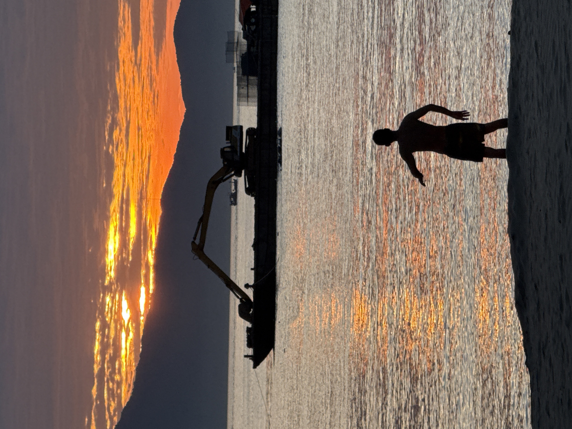 This vertical photograph captures a sunset over a body of water with a mountainous backdrop, consistent with coastal areas near Đà Nẵng, Vietnam. The sky displays a progression of orange and yellow hues, with direct sunlight visible through horizontal cloud formations above a range of dark, silhouetted mountains that define the distant horizon.
In the mid-ground, a dark, flat-deck barge floats on the water. A large hydraulic excavator, also in silhouette, is positioned on the barge with its boom extended upwards and to the left. Further in the background, indistinct silhouettes of additional structures or vessels are present on the water to the right.
The surface of the water reflects the warm tones of the sky, creating shimmering patterns. In the foreground, a dark, sandy area transitions into shallow water where a silhouetted human figure stands, facing away from the camera. The individual, appearing male and dressed in shorts, has one arm slightly raised and extended to their left.