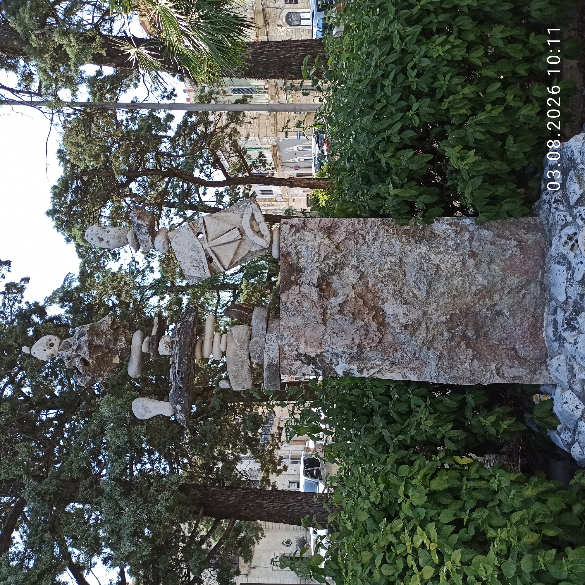 An outdoor scene depicts a stone sculpture composed of various balanced rocks of different sizes and shapes, standing amidst lush green bushes and trees. The sculpture is positioned in the foreground, with buildings and more trees visible in the background. The sky is bright and clear. The sculpture appears to be abstract, with some rocks resembling figures or faces. There are no people or animals present in the immediate vicinity of the sculpture. The date stamp "03.08.2026 10:11" is visible in the lower right corner of the image. The location is identified as Ruvo di Puglia, Italy.