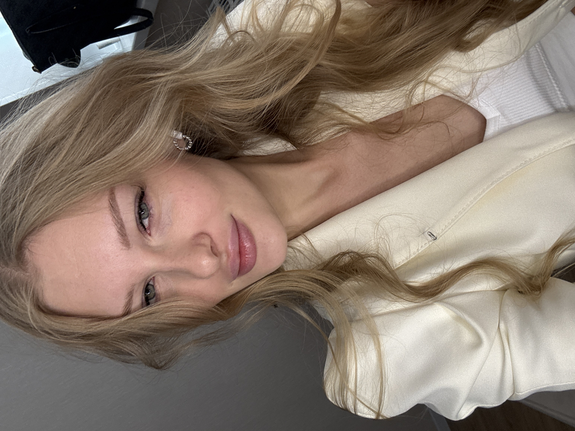 A close-up, medium shot captures a young Caucasian woman with long, wavy blonde hair from a slightly high angle. She is wearing a cream-colored satin blazer over a white ribbed tank top and a small, circular, sparkling earring in her left ear. Her makeup includes eyeshadow and mascara, and her lips are glossed. Her gaze is directed towards the viewer with a subtle smile. The background is out of focus, appearing as a gray wall and a black bag in the upper right corner. No specific location details for Perm, Russia are discernible.