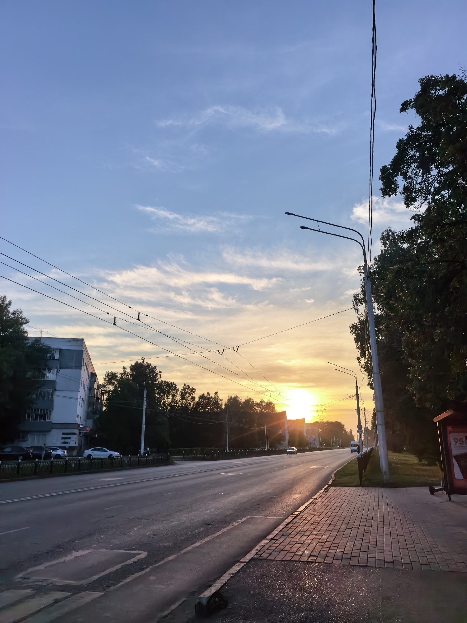 The image depicts an urban street scene at sunset in Sterlitamak, Russia. The sun is setting on the horizon, casting a warm orange and yellow glow across the sky and reflecting on the wet asphalt of the road. The sky is a gradient of blue and soft clouds, with wispy streaks indicating movement. Overhead, a complex network of power lines and trolley wires crisscrosses the sky, supported by utility poles.
To the left of the road, a multi-story apartment building with a grey and white facade is visible, its windows reflecting the sky. Several cars are parked along the side of the road and on the street. Trees line the right side of the road, forming a dense canopy that partially obscures the sky and buildings.
The sidewalk in the foreground is paved with reddish-brown bricks and appears slightly damp, further enhancing the sunset reflections. A bus stop shelter with a red and white advertisement panel is visible on the far right. The road itself shows signs of wear and tear, with some patched areas and faded lane markings. The overall atmosphere is tranquil, with the sunset creating a sense of calm over the urban environment. There are no individuals clearly visible or engaged in any discernible actions.