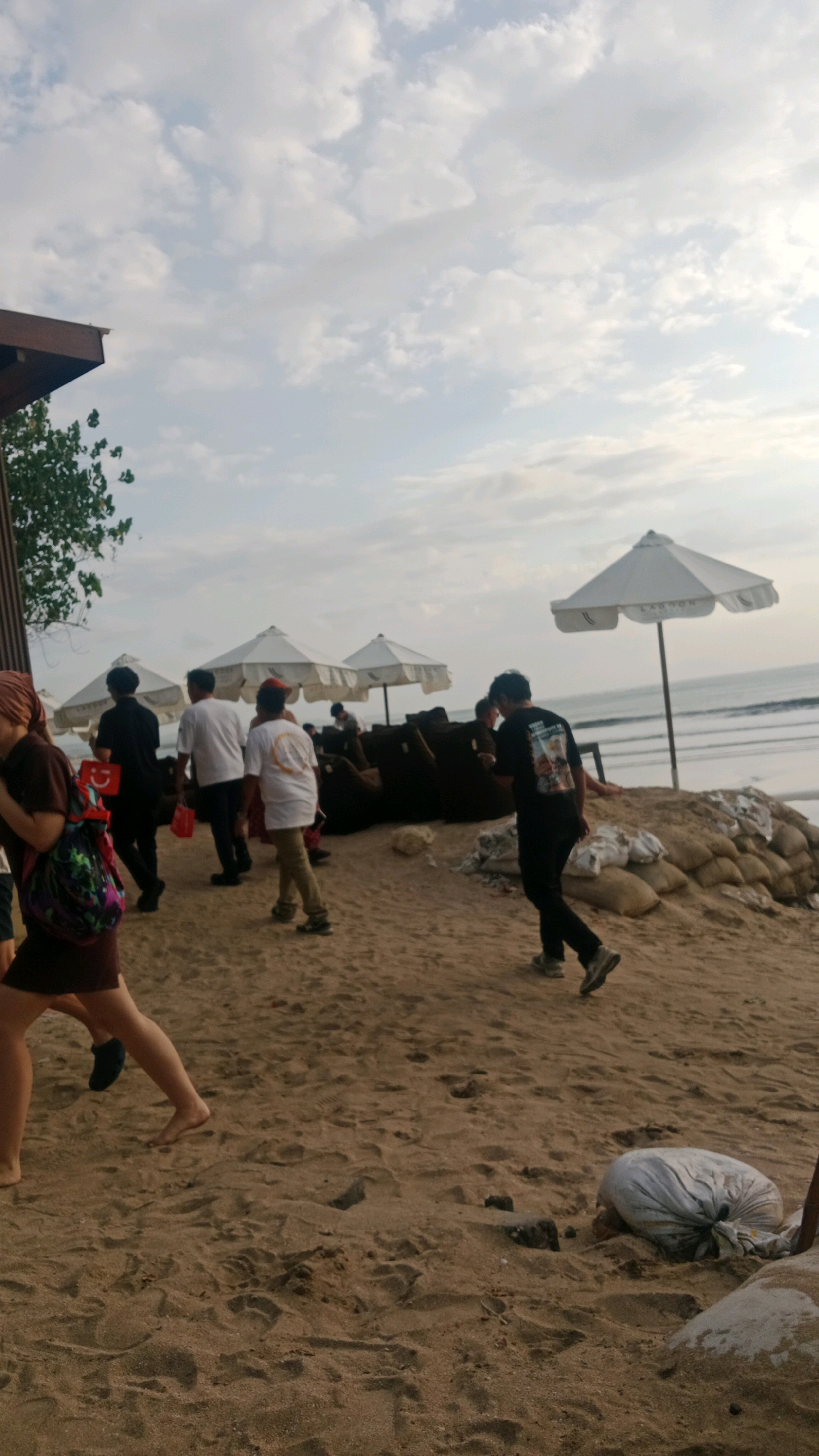 A group of people are walking on a sandy beach in Kuta, Indonesia, near the ocean. The sky is cloudy. There are white umbrellas and beanbag chairs on the sand, suggesting a resort or beach club. Some of the people are carrying bags. The sand is marked with footprints, and there are some bags of sand piled up. The overall scene is casual and relaxed, with people enjoying the beach environment.