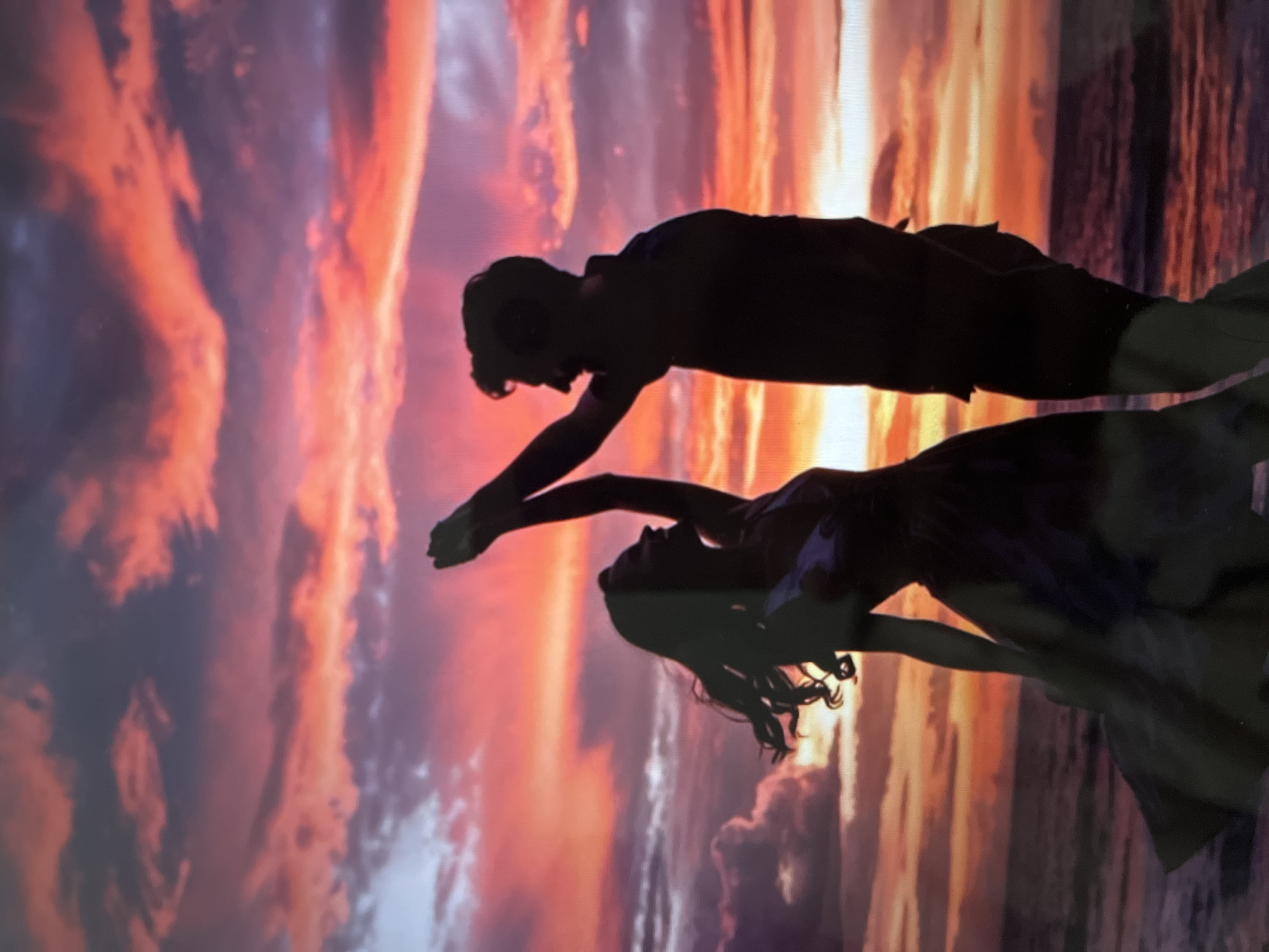 An ethereal silhouette of a couple dancing on a beach against a vibrant sunset is captured. The sky explodes with fiery oranges, pinks, and purples, with dramatic clouds streaking across the horizon. The couple, rendered in dark silhouette, are positioned centrally, with the woman on the left and the man on the right. The woman is facing the man, her arms raised and extended, her hair flowing outwards. The man is also facing her, his arms raised as if leading the dance. Their bodies are entwined in a graceful pose, suggesting a moment of romantic connection. The foreground features the gentle waves of the ocean lapping at the shore, with the dark sand providing a stark contrast to the luminous sky. The overall impression is one of romance, beauty, and the magic of a sunset.