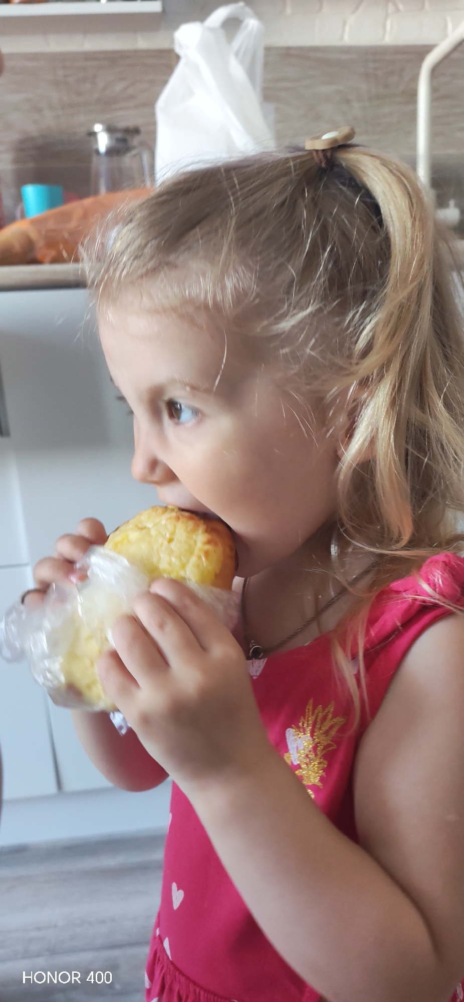 A young girl with blonde hair, styled in a high ponytail, is eating a baked item. She is wearing a red dress with white heart patterns and a gold design on the chest. The baked item is wrapped in clear plastic. The background includes a kitchen counter with various objects and a white wall. The location is not specified in the image or context, it is assumed it takes place in Noviy, Russia. "HONOR 400" is printed on the bottom of the image.