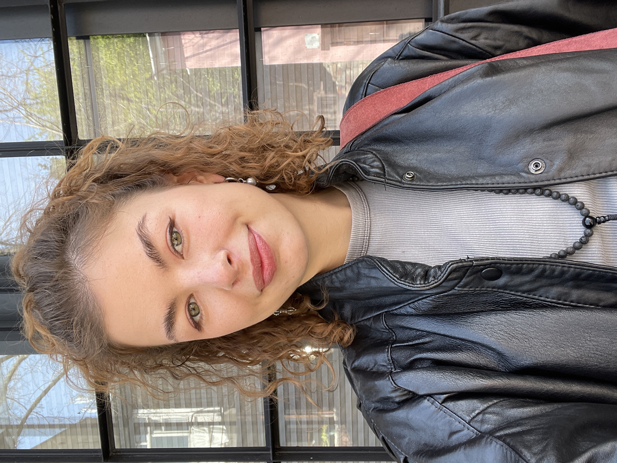 A young woman with light skin, green eyes, and voluminous, light brown curly hair is depicted in a portrait-style photograph. She looks directly at the camera with a subtle, pleasant smile and a calm expression. She wears a light grey ribbed top, covered by a black leather jacket, which has a reddish-pink strap or lining visible over her right shoulder. A black beaded necklace and small, possibly silver or pearl, earrings accessorize her outfit.
The woman is positioned indoors, likely seated or standing, in front of a large window with dark frames. Through the window, an outdoor scene with trees showcasing early green foliage is visible, suggesting a springtime setting. The natural light indicates it is daytime. The image has a tilted orientation, with the woman's head appearing to lean to her left. There is no visible text in the image. The location context for the photo is Tbilisi, Georgia.