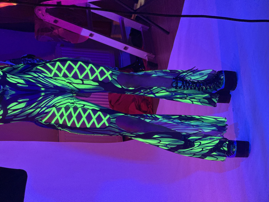The image displays the lower torso and legs of an individual standing in an indoor environment under ultraviolet (UV) illumination. The subject is clad in a dark, form-fitting garment featuring intricate, bioluminescent-like neon green patterns. The garment incorporates cut-outs on the upper thighs, detailed with glowing neon green crisscross lacing, and expands into flared bell-bottoms at the ankles. Black, high-platform, lace-up boots are worn on the feet. The scene is bathed in a predominant purple and blue ambient light, causing the neon green elements of the attire to fluoresce brightly. In the background, a light-colored backdrop is visible to the left. To the right, a silver stepladder stands, with a red object and a white radiator visible behind it against a wall. The floor is light-toned, reflecting the UV light. This setting is consistent with a studio or event space located in Sofia, Bulgaria. The specific address or venue within Sofia is not ascertainable from the visual evidence.
