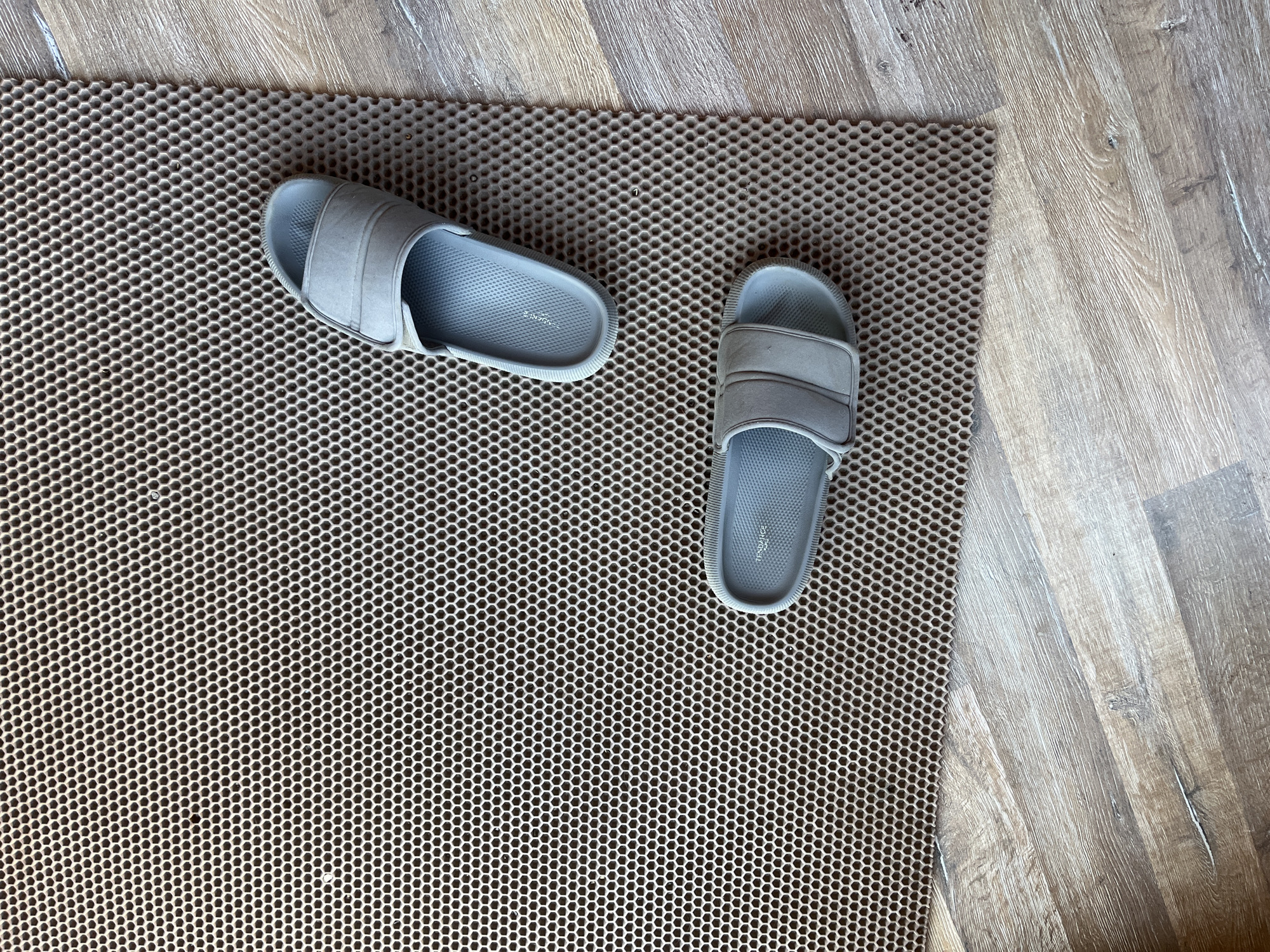 Two light grey slides with two textured straps are placed on a brown honeycomb-patterned mat. The mat is situated on a wooden floor with a light brown, grey, and white wood grain pattern. The image is taken from a top-down perspective. There are no people or discernible locations in Saratov, Russia.