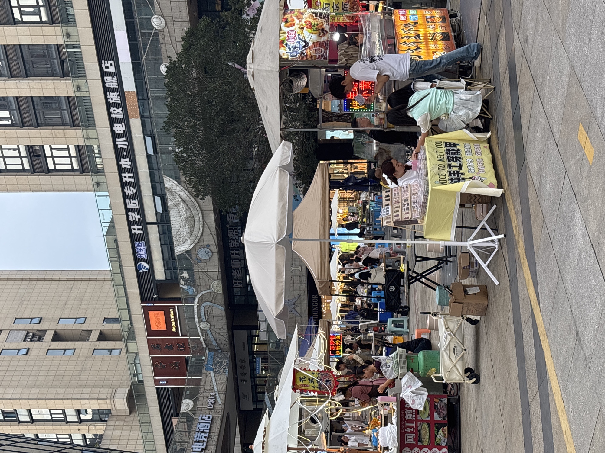 Here's a detailed description of the image:
**Overall Impression:**
The image captures a bustling outdoor market scene. Several stalls are set up along a pedestrian walkway, each covered by white umbrellas. The stalls are lined with various vendors and customers. A tall building with glass windows and signage is visible in the background.
**Specific details:**
* **Stalls:** The stalls are selling a variety of items, including jewelry, food, and potentially other crafts. Each is lit by a light. The food stall is selling noodles, fried rice and fried noodles. One stall displays a sign that reads "NICE TO MEET YOU" and advertises handmade products.
* **People:** People are both selling items from their stalls and purchasing them from other stalls.
* **Building:** In the background is a multi-story building with glass windows. The building also has the names "RepairMan," "GAUERANG," "YOUNGOR," and "升学匠专升本水电校旗舰店," and "好老师升学潮专" on it.
* **Ground:** The ground is paved with light-colored paving stones. A yellow line is painted on the ground.
* **Environment:** The market is set in an urban environment. Trees are partially visible in the background.
* **Signs:** There are signs with Chinese characters indicating the names of businesses or product descriptions.
**Location Identification (based on the signage):**
Based on the signage in the background, the image could be in Yongchuan, China.