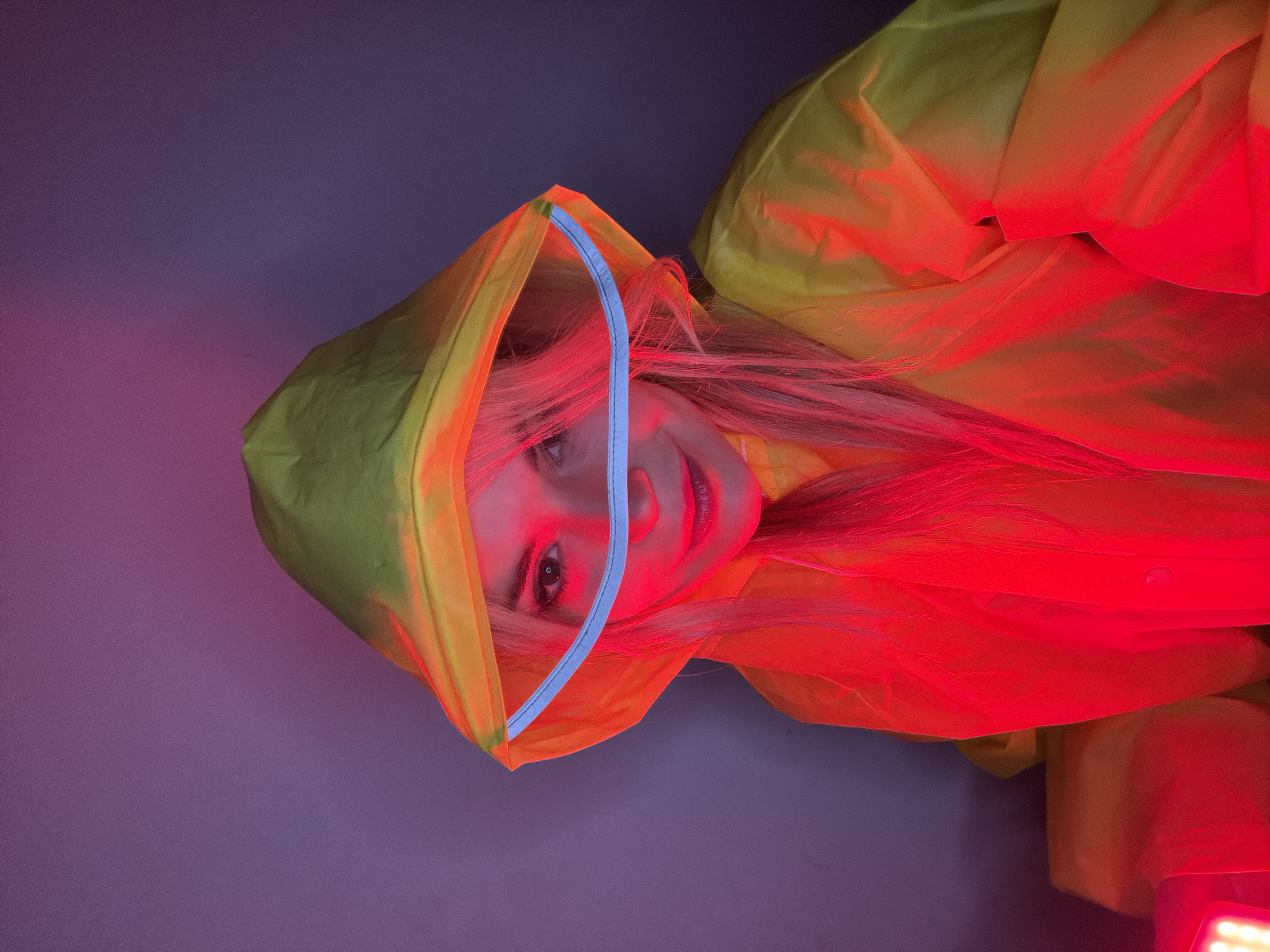 The image portrays a young woman in a vibrant, artificially lit setting, possibly a studio, in Budva, Montenegro. She is wearing a bright yellow-green raincoat with the hood up, featuring a clear plastic brim outlined in blue. The image is oriented with her head towards the left side of the frame, and she is looking upwards, directly at the viewer, with a gentle smile on her face.
A strong red light dramatically illuminates the right side of the scene, casting intense red and orange tones across her face, her long blonde hair, and the right portion of her raincoat. This intense lighting creates deep shadows and highlights, making the raincoat appear to shift colors. The background behind her gradually transitions from purple on the left to darker blueish-purple shades on the upper right, contributing to the surreal and colorful atmosphere. The combination of the raincoat and the striking lighting suggests a stylized photoshoot or artistic portrayal. There is no visible text in the image.
