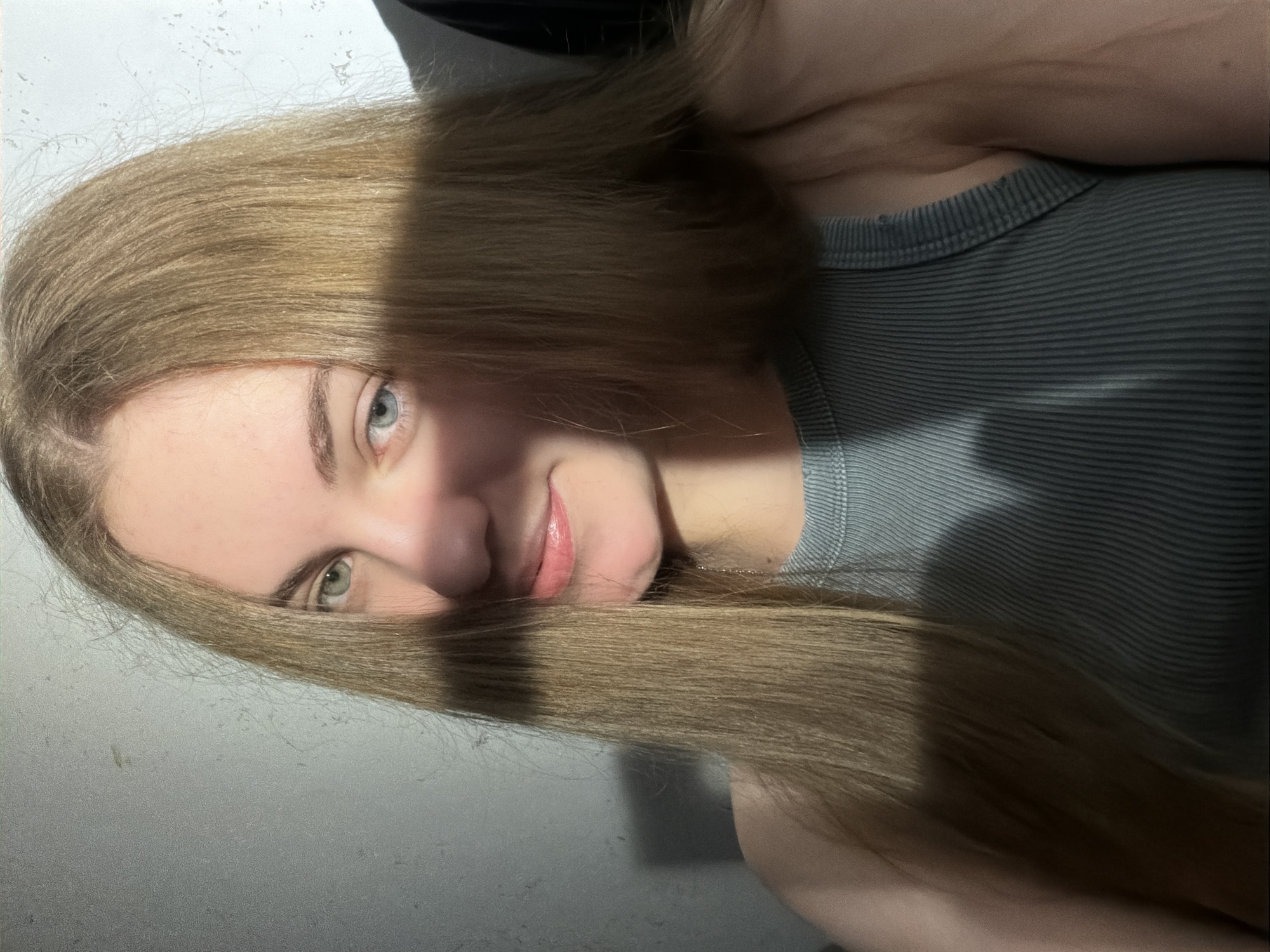 A young person with long, blonde hair is taking a selfie. The image is rotated 90 degrees counter-clockwise, so the person appears to be lying down. They are wearing a grey, ribbed tank top. Their eyes are light blue and green, and they have a slight smile on their lips, which are painted with a peachy-pink lipstick. The background is a plain, textured wall, and there is a shadow cast across the person's face and hair, suggesting indoor lighting or a strong light source from the side. The overall mood is casual and intimate. There is no visible text in the image.