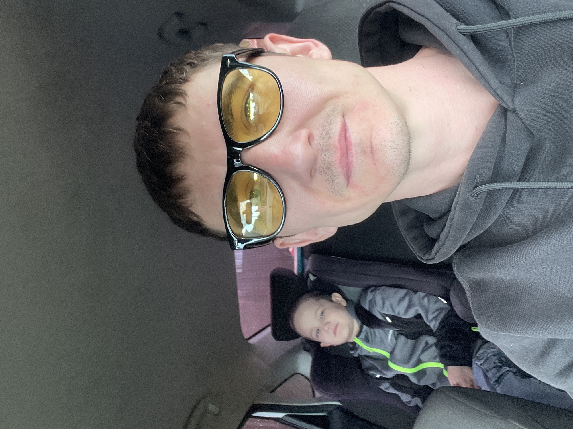 A man wearing yellow-tinted sunglasses is taking a selfie in a car. He is positioned in the front seat, and the camera is angled to capture his face and upper body. He is wearing a dark gray hoodie. In the background, a young child is visible in a car seat. The child is wearing a gray and black jacket with a neon green stripe. The child is looking towards the camera with a neutral expression. The setting appears to be inside a car, likely during the daytime, given the natural light illuminating the subjects. The overall mood is casual and relaxed. There is no visible text in the image.