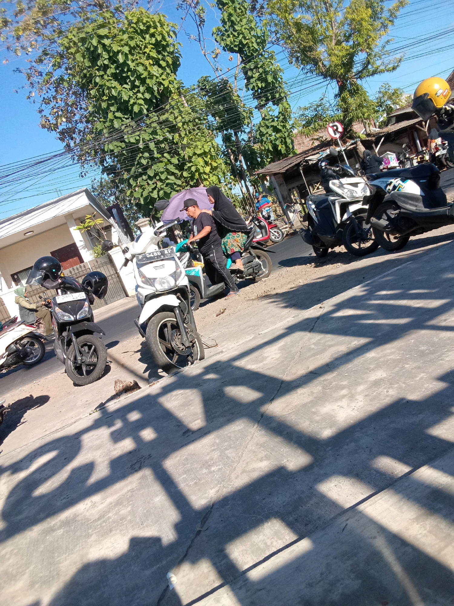 This is a ground-level, slightly angled, medium shot of a street scene in Jember, Indonesia, on a sunny day. The foreground is dominated by a concrete road surface casting long, angular shadows of trees and possibly a fence or structure. In the mid-ground, several motorcycles are parked or in motion. Two black scooters are prominent on the left, each with helmets on their handlebars. A white scooter with license plate "P 5486 RF" is positioned centrally. Further back, a white scooter with blue accents and a yellow helmet on its handlebars is visible. To the right, a dark-colored motorcycle with patterned decals is parked. Behind the motorcycles, there are low-lying buildings with corrugated roofs and a dense green tree canopy. A few people are present, including individuals on or near the motorcycles, some wearing headscarves and casual clothing. A "No Parking" sign is visible on a pole in the background. The overall impression is a typical roadside scene in a developed urban or suburban area.