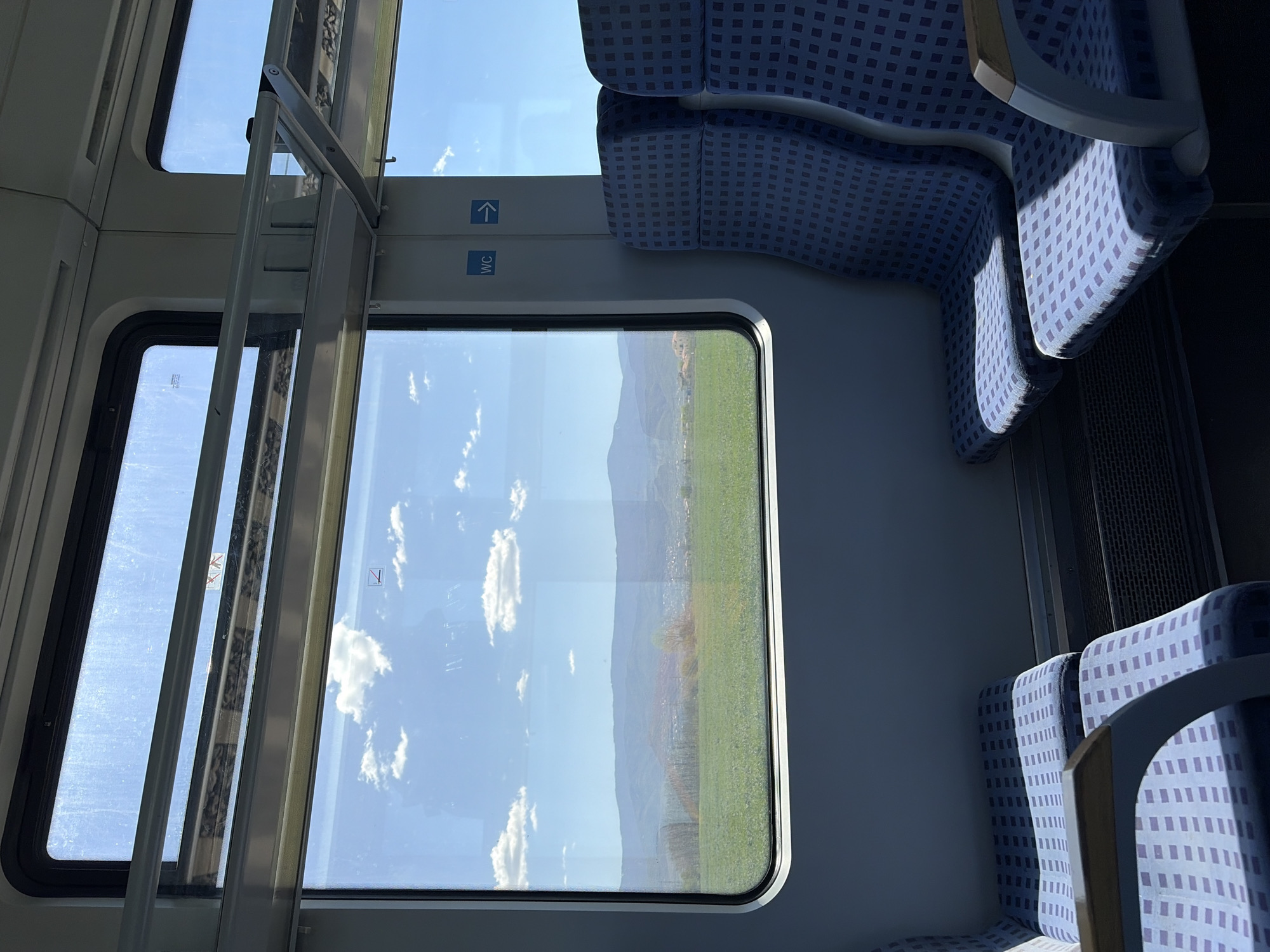 The interior of a passenger train is visible through a partially open window. The window frames a view of a landscape with green fields transitioning into mountains under a clear blue sky dotted with white clouds. The train's interior is marked by blue patterned seats, and signage indicating the location of the restroom. The image is shot from an eye-level perspective inside the train.