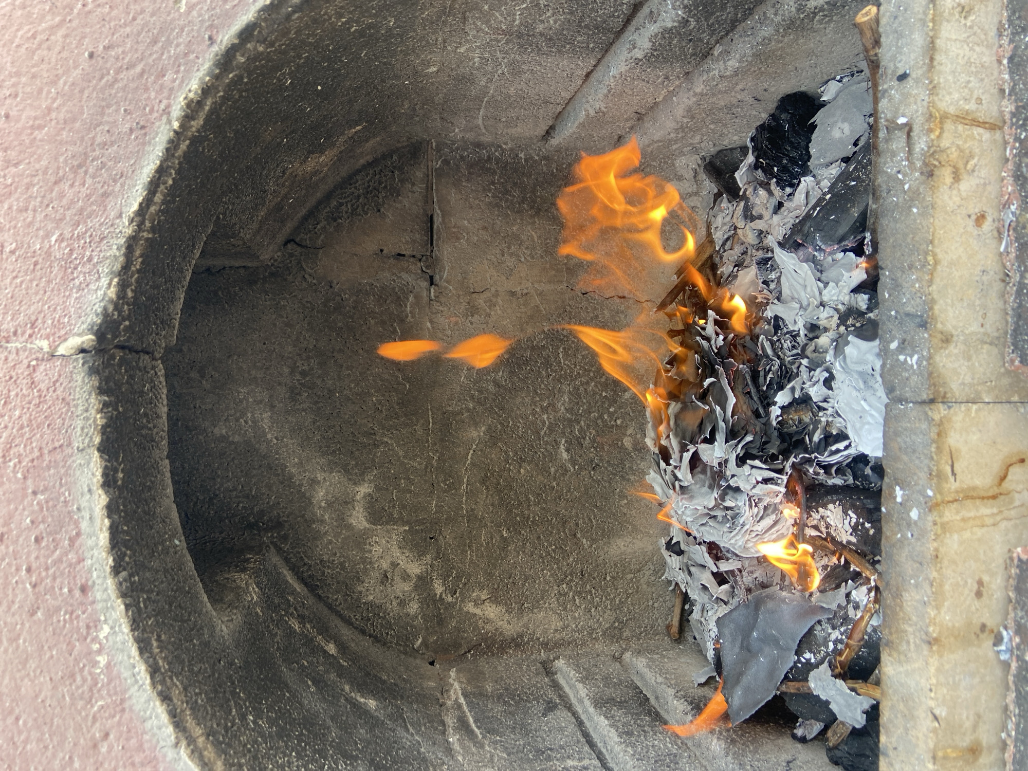 A fire burns within a gray brick or concrete structure. The flames are orange and yellow and are consuming crumpled, white and gray paper or material. The structure's interior appears to be aging or weathered, with cracks visible in the surface. The outside of the structure appears to be coated in a light pink coating. It is unknown if this image was taken in Budva, Montenegro, as it is not visible within the image.