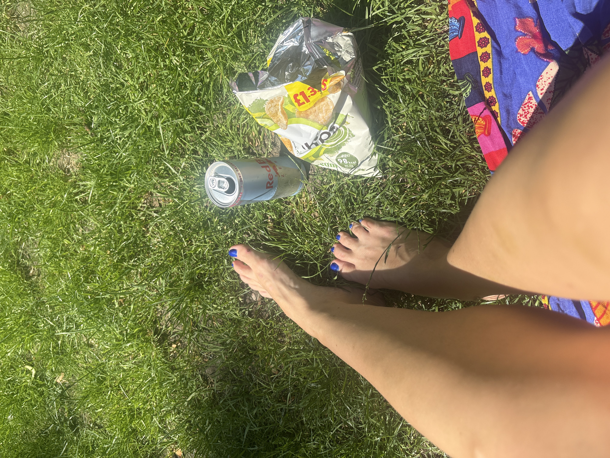 This media file depicts a person's legs and feet resting on bright green grass. The individual's toenails are painted a vibrant blue. In the scene, there is an open bag of crisps and a Red Bull can. A colorful, patterned fabric, possibly a blanket or clothing item, is visible to the right of the frame. The lighting suggests it is daytime, with bright sunlight casting shadows on the grass, indicating a sunny day. The presence of grass and outdoor items implies an outdoor setting, likely a park or garden. There is text visible on the crisp bag, including "£1.39" and nutritional information stating "98 kcal per serving." The Red Bull can also has visible text, including the brand name "Red Bull." The overall impression is one of relaxation and enjoying a snack outdoors on a sunny day.