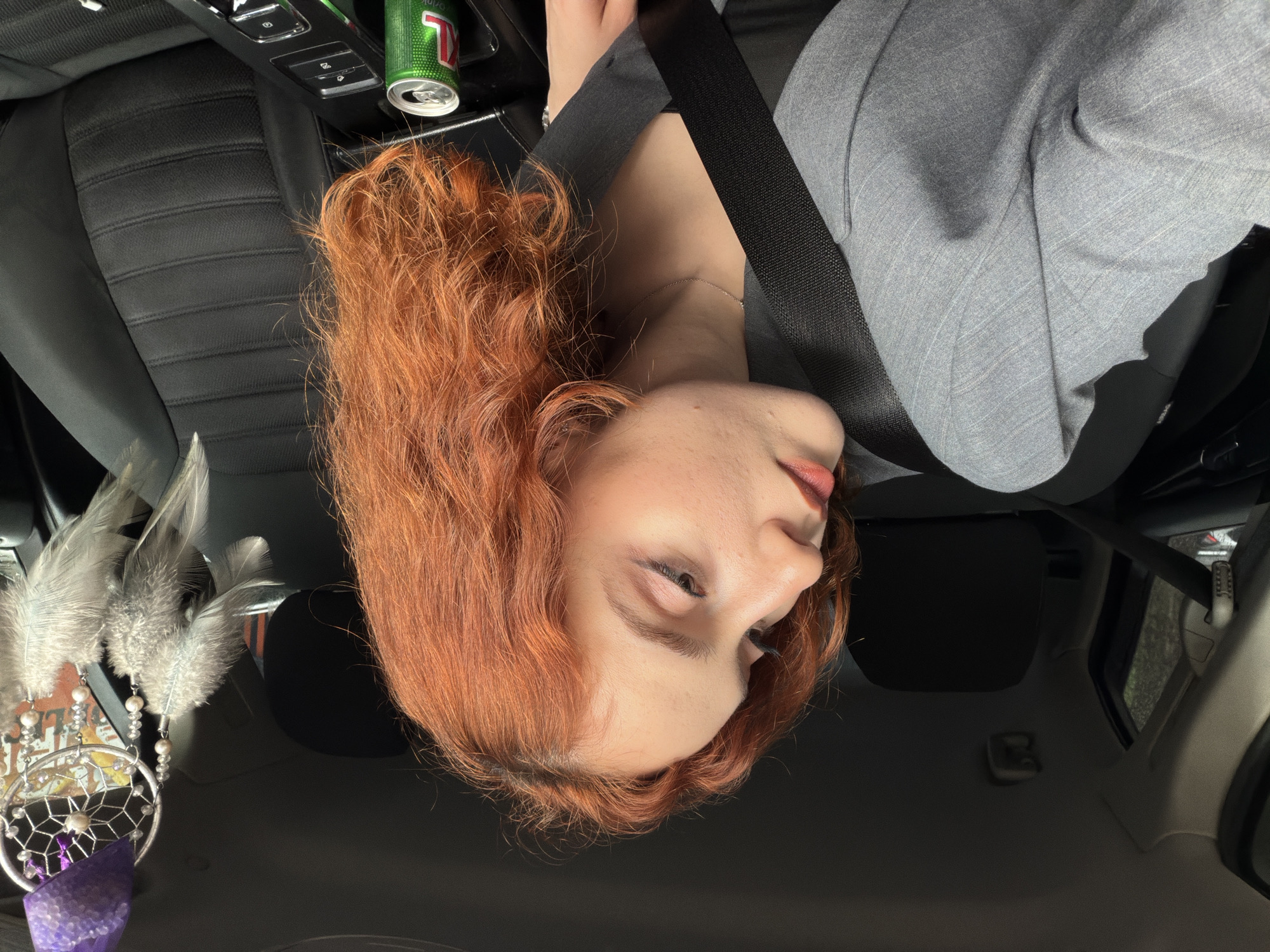 A woman with auburn curly hair, wearing a gray suit jacket and a seatbelt, sits in the driver's seat of a car. A partially consumed can of "Xtra Drink" is in the center console. A dreamcatcher with feathers and purple stone hangs from the rearview mirror. The environment appears to be the interior of a car, but there is no evidence in the image to ascertain that the location is Tbilisi, Georgia.