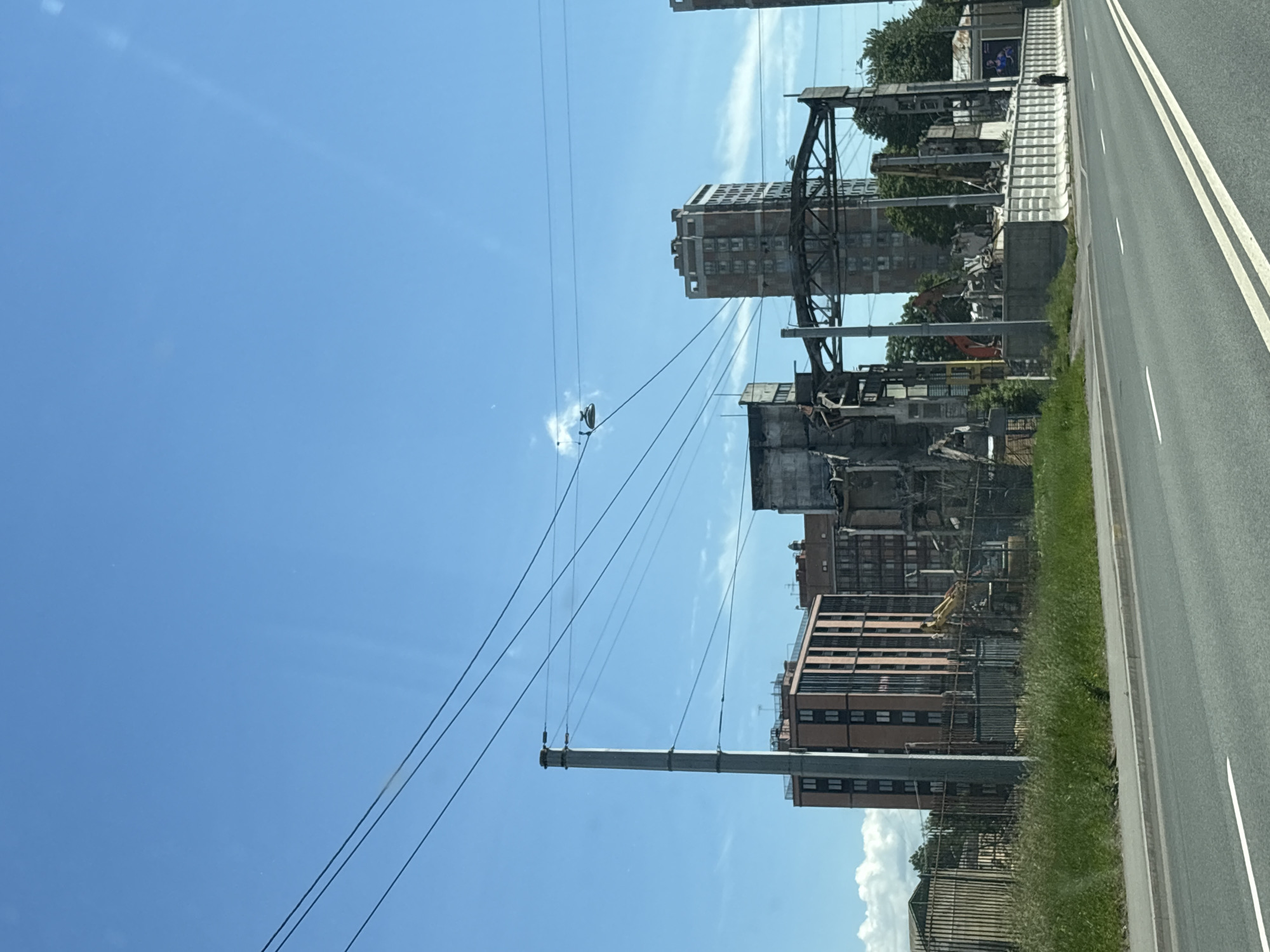 The image captures a street view in Saint Petersburg, Russia, on a clear day with a blue sky. The foreground shows a paved road with white lane markings and a strip of green grass and vegetation bordering it. Beyond the grass, a fence encloses a construction or demolition site. Various buildings are visible, including a tall, modern building with orange-pink cladding and dark windows, and other structures that appear to be under demolition, with exposed frameworks and debris. Construction equipment, such as an excavator, is present at the site. Overhead, power lines stretch across the frame, supported by a tall utility pole on the left. In the distance, several multi-story buildings, including a reddish-brown tower, are visible. A single person is walking along a sidewalk in the mid-ground, to the right of the demolition site. The overall scene suggests urban development or redevelopment in progress.