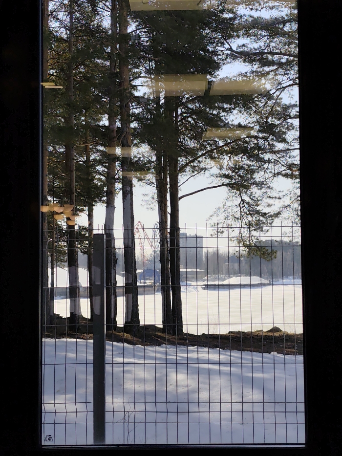 The image provides a forensic view through a window onto a winter landscape in Chaikovsky, Russia. Dark vertical borders frame the scene, with multiple reflections visible on the glass, including several horizontal rectangular light strips across the upper portion and a cluster of small, warm-toned, semi-circular shapes along the mid-left edge.
In the immediate foreground, a grey wire mesh fence spans the lower and central sections of the view, featuring a distinct dark vertical fence post positioned in the lower-left quadrant. Immediately behind the fence, snow covers the ground. The midground is marked by a sparse line of tall pine trees, several of which display white-painted bands on their lower trunks. Beyond these trees lies a broad expanse of frozen, snow-covered water, identified as part of the Kama River or Votkinsk Reservoir.
On the far bank of the frozen water, a prominent red steel structure is visible, identifiable as a component of the "Snezhinka" ski jumping complex. To its right, a multi-story building with a light-colored facade stands. The sky above is pale blue, indicating daylight conditions with no significant cloud cover. No individuals are present, and no actions or interactions are depicted within the scene.