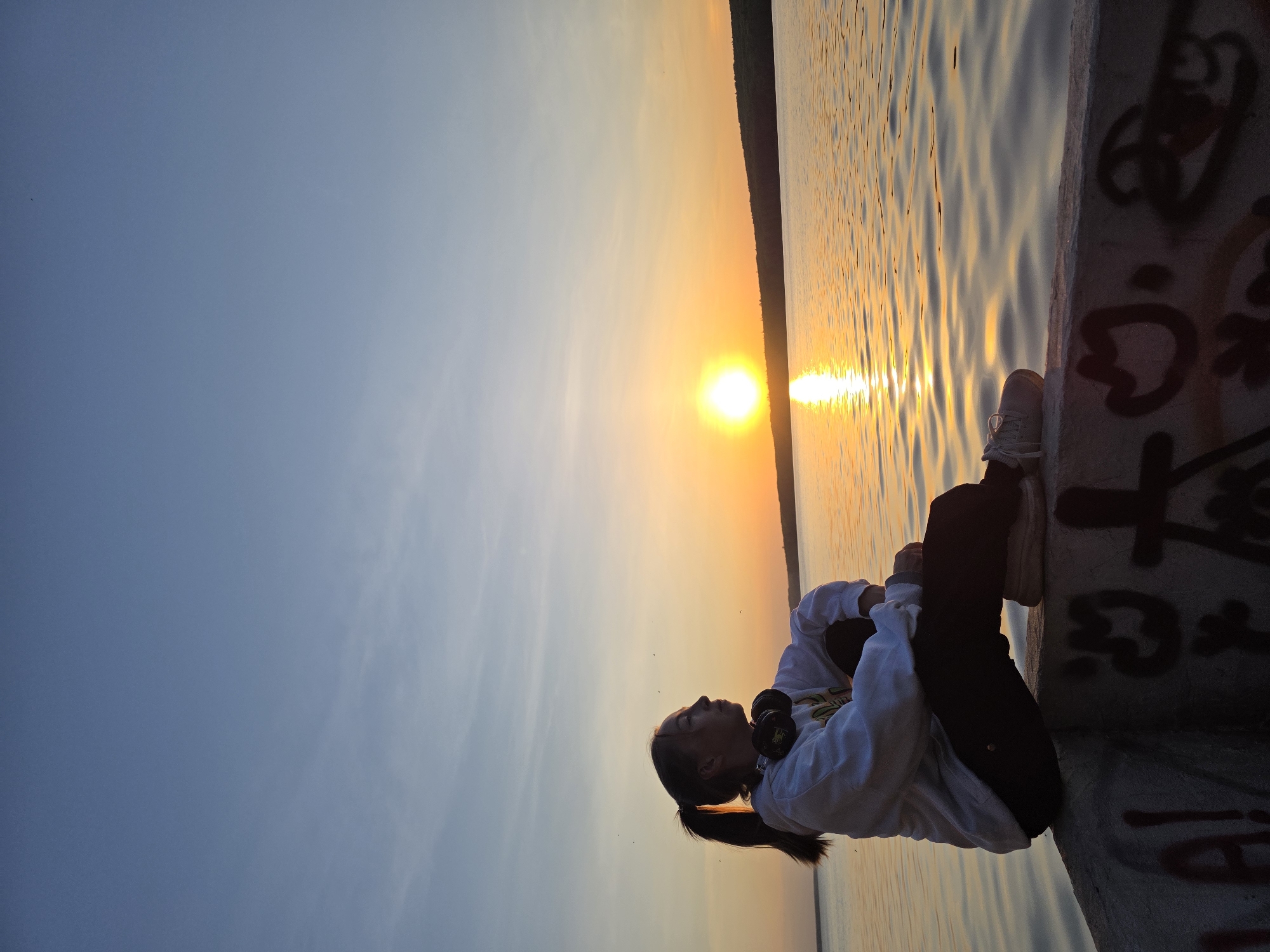 A young woman with a ponytail is sitting on a concrete ledge, facing left towards the sunset, with a body of water behind her. She wears a white sweatshirt with headphones around her neck and dark pants. The concrete ledge she is sitting on appears to have graffiti on its side. The location is likely the city of Chaikovsky, Russia, as specified in the prompt. The sun is low on the horizon, reflecting golden light on the water. The sky is a gradient from light blue to orange near the horizon.