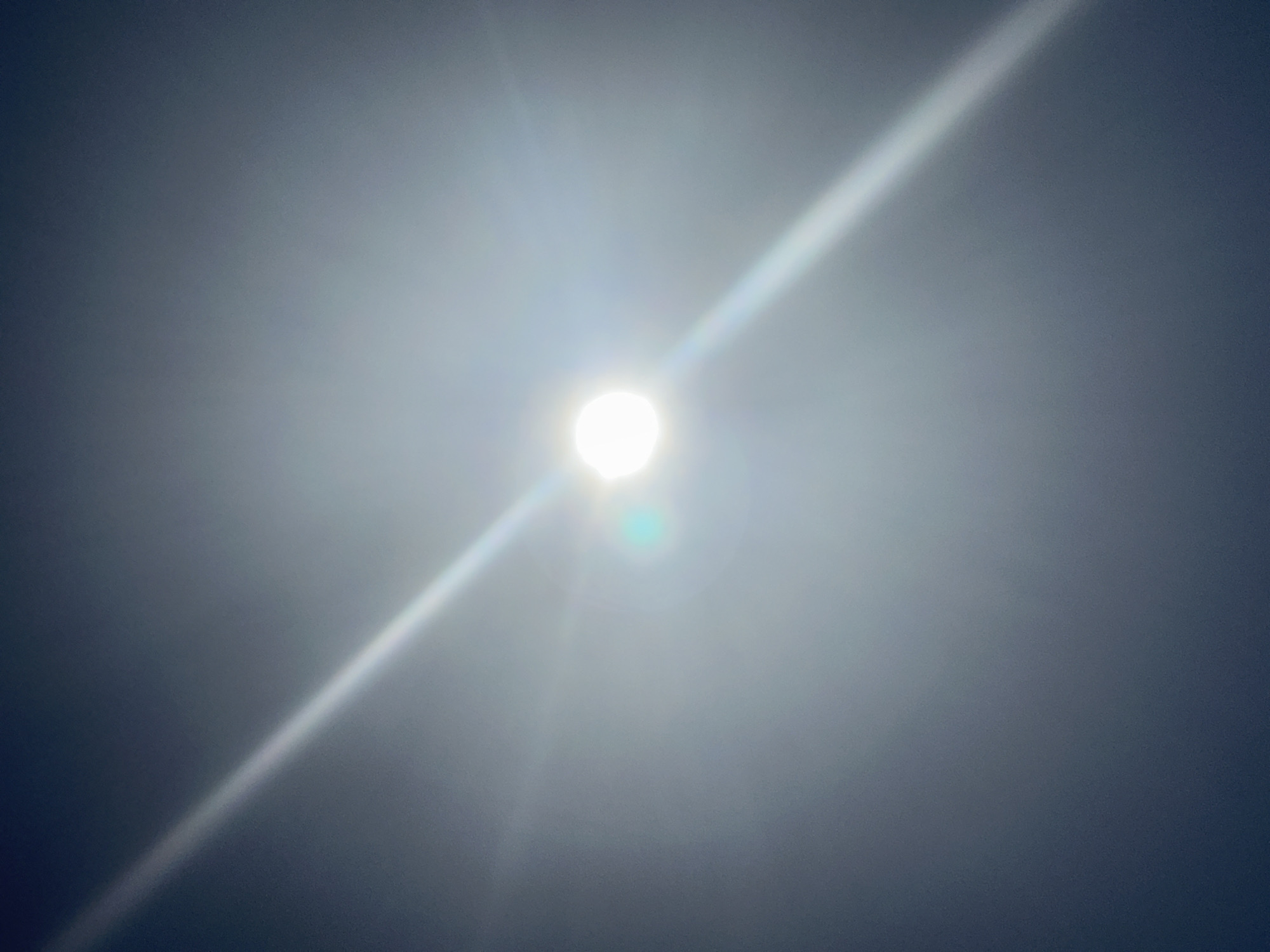 The image shows a bright light source, likely the sun, obscured by haze or thin clouds. A diagonal streak of light emanates from the central point. The background is a muted grey-blue, suggesting the sky. The location is Unknown, France.