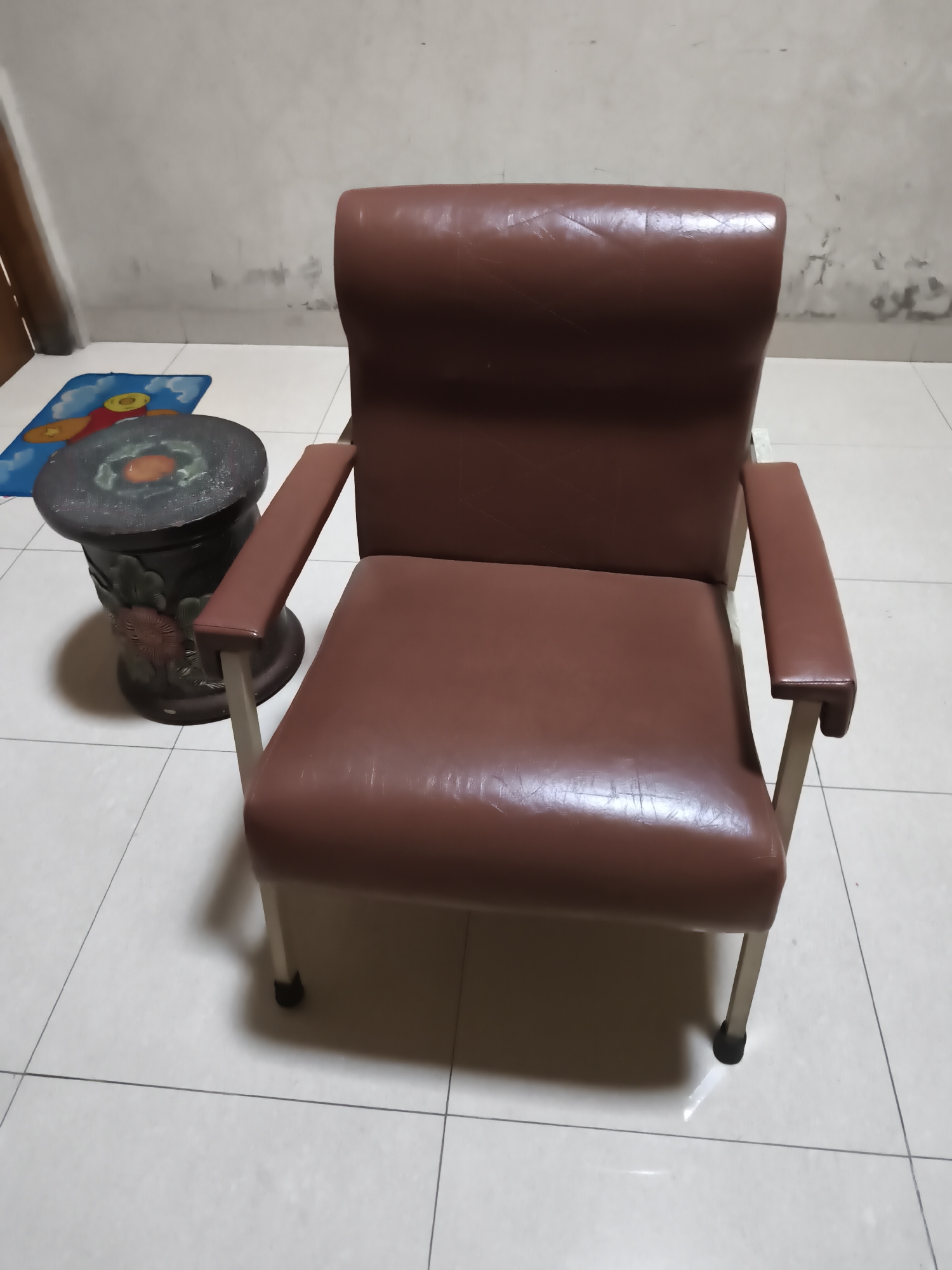 A brown leather armchair is positioned on a tiled floor in front of a textured, light-colored wall. To the left of the armchair, a small, dark, cylindrical stool with decorative carvings is visible. A colorful mat with an image of a plane is placed on the floor near the stool. The lighting in the scene suggests an indoor environment, likely a room or hallway.