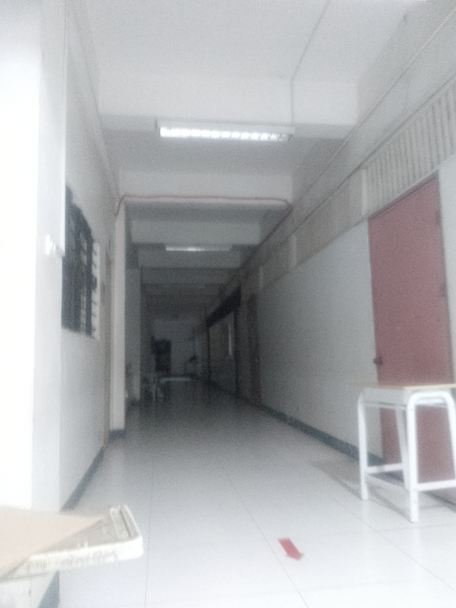 The image depicts a dimly lit hallway, likely indoors, characterized by light-colored tile flooring and neutral-toned walls. Overhead fluorescent lighting fixtures provide illumination, though the area appears somewhat dark. A row of windows is visible on the left side of the hallway. A light red door stands on the right side of the hallway along with a stool, and a small, red, arrow-shaped sticker is on the floor. The environment suggests a public or institutional building. The city of Davao City, Philippines, cannot be determined.