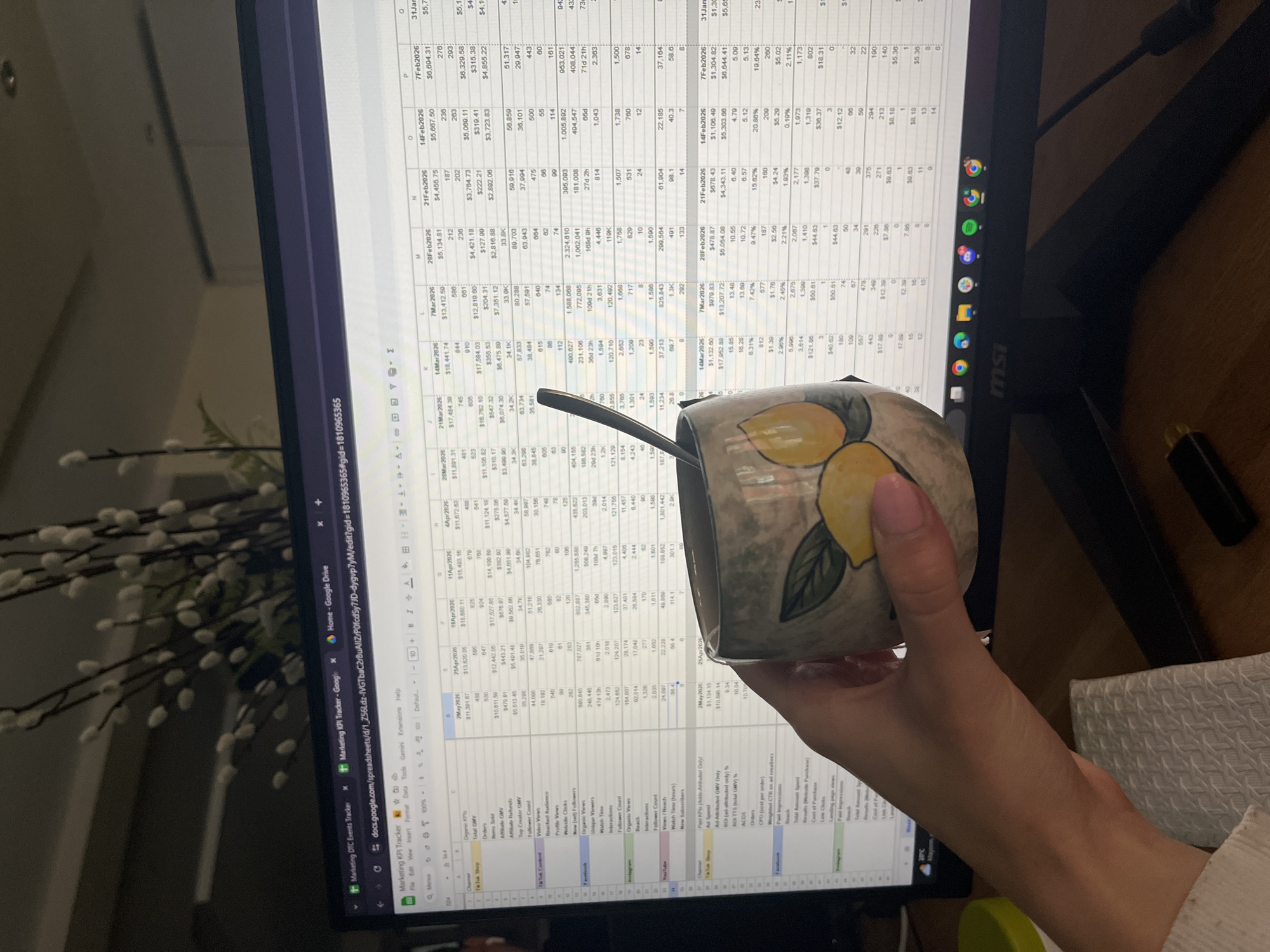 A person is holding a lemon-patterned mug in front of a computer screen displaying a spreadsheet titled "Marketing KPI Tracker" in Google Sheets. The spreadsheet contains data organized by dates ranging from 2 May 2026 to 31 January 2026, with rows representing various metrics like "Total GMV," "Orders," "Items Sold," and "Organic GMV." The screen also displays open tabs for "Marketing DTC Events Tracker" and "Home - Google Drive." The computer is an MSI-branded monitor on a desk. A decorative plant with white blooms is partially visible in the background to the left of the monitor. A white patterned object is on the right side of the desk. A green object is partially visible under the monitor.