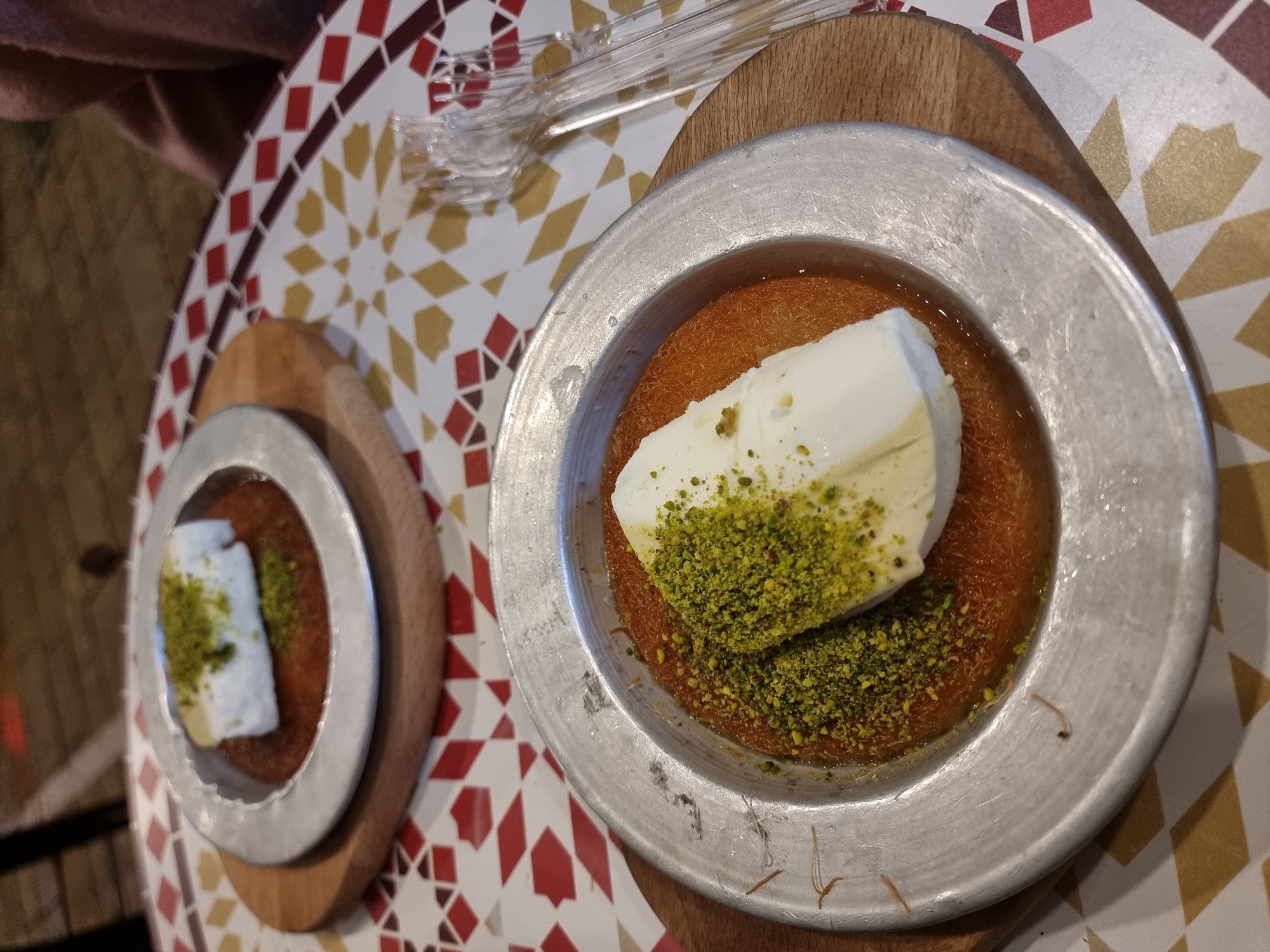 The image depicts two plates of what appears to be Kunafa, a Middle Eastern dessert, topped with white cheese or ice cream and chopped pistachios. The desserts are served on metal plates placed on small wooden boards. The plates rest on a table with a geometric tile pattern in red, white, and gold. In the background, there are clear plastic forks. Given the dessert and the table's style, it's plausible the location is in Tel-Aviv, Israel, a city known for its diverse cuisine that includes Middle Eastern and Mediterranean influences. However, without further information or visual cues, it's impossible to definitively confirm the specific location within Tel-Aviv. There are no people visible in the frame. The scene focuses on the presentation of the food.