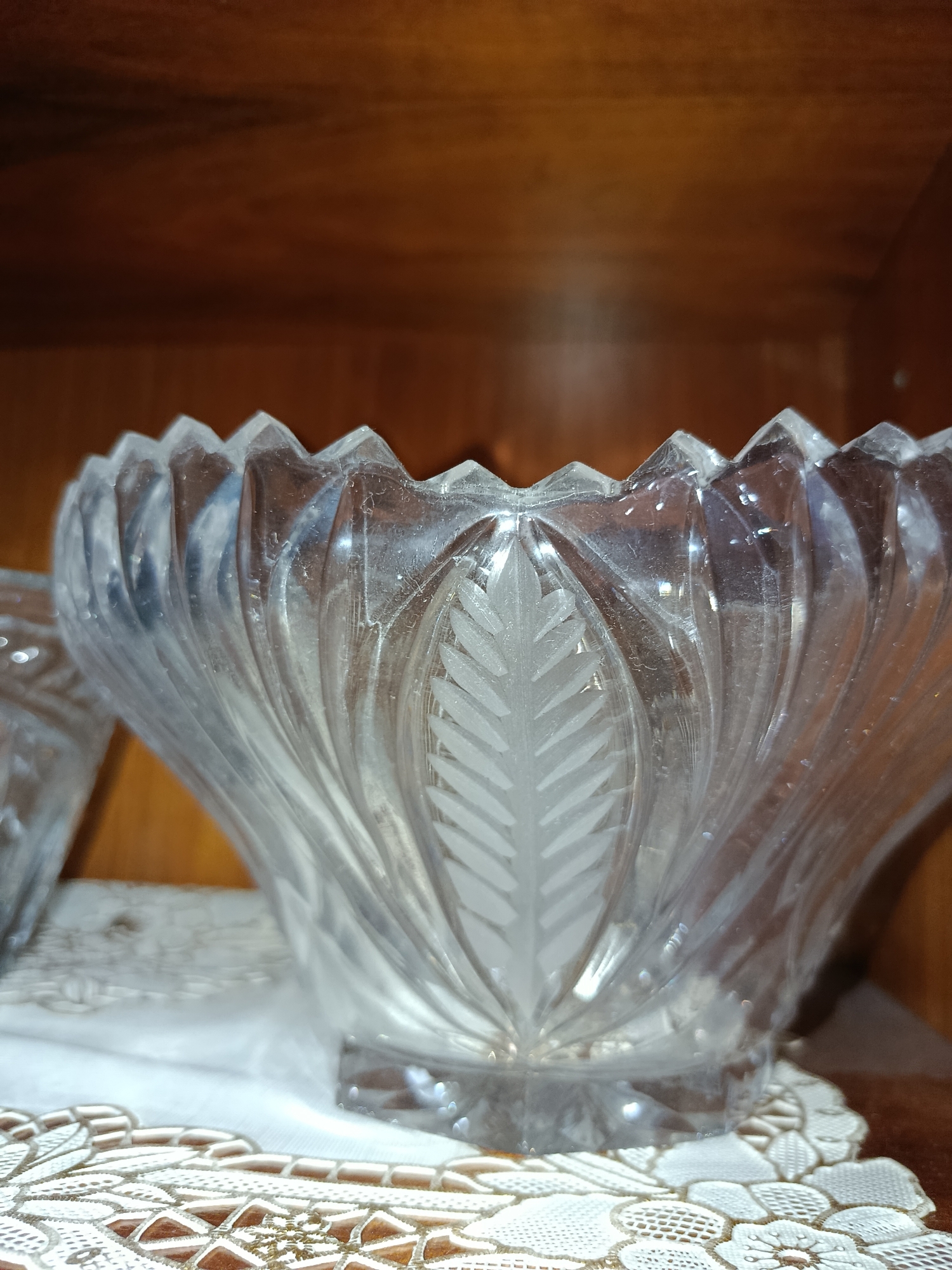 A close-up, eye-level shot shows a clear glass bowl with etched floral patterns, placed on a doily inside a wooden cabinet. The bowl has a fluted rim and a raised base. The doily is white with intricate lace-like patterns. The background is a wooden surface. The scene appears to be indoors, possibly in a home or display cabinet. There is no indication of the location being in Tashkent, Uzbekistan.