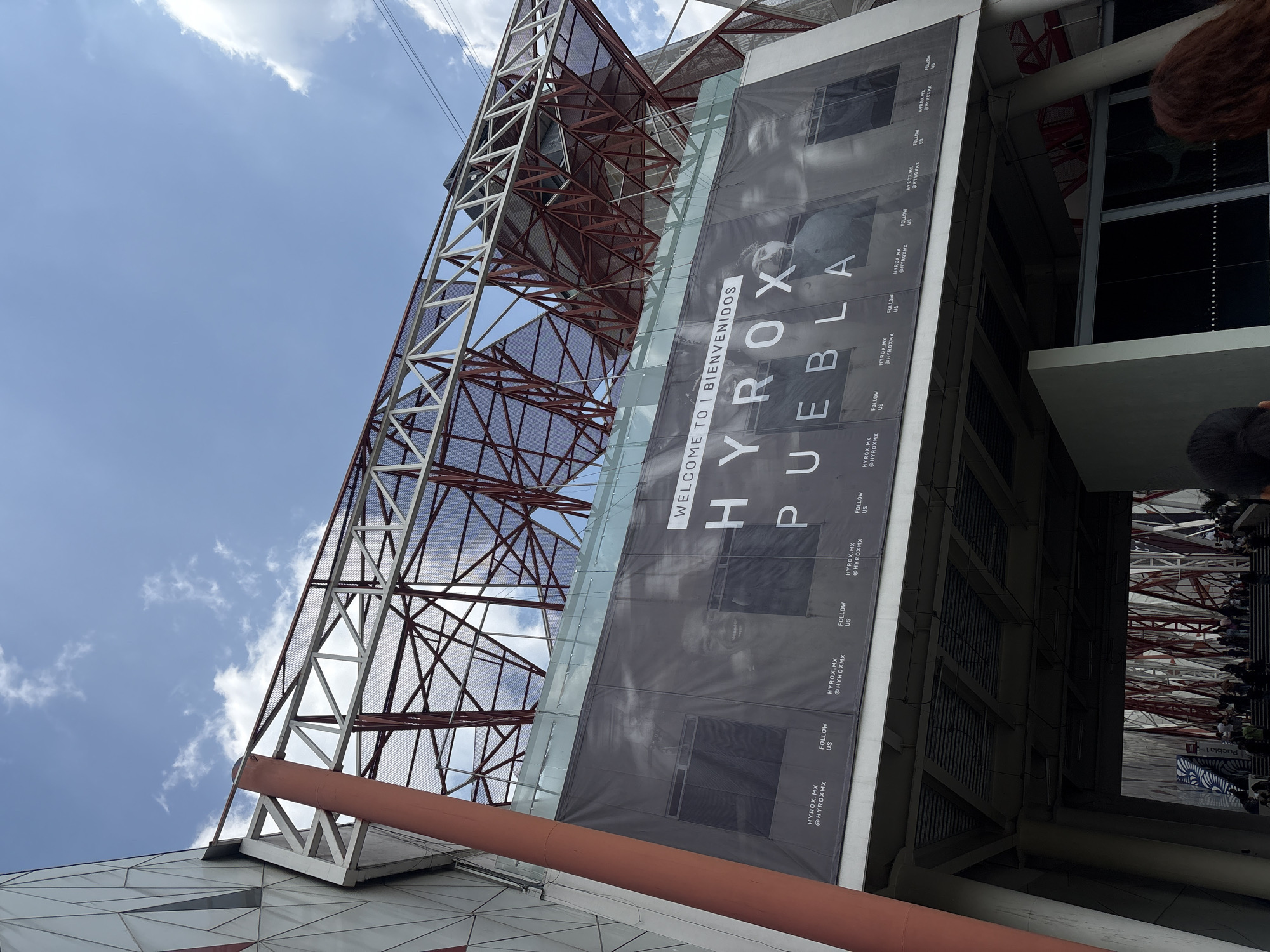 The image depicts a low-angle view of the Hyrox Puebla building in Puebla, Mexico. A large banner on the building reads "WELCOME TO | BIENVENIDOS HYROX PUEBLA" in large, vertical lettering. Smaller text on the banner includes "HYROX.MX" and "@HYROXMX" with the phrase "FOLLOW US" repeated multiple times. The building features a steel support structure with a red-brown and silver finish. The sky is visible behind the structure, showing a partly cloudy day. People are visible inside of the building at the entrance.