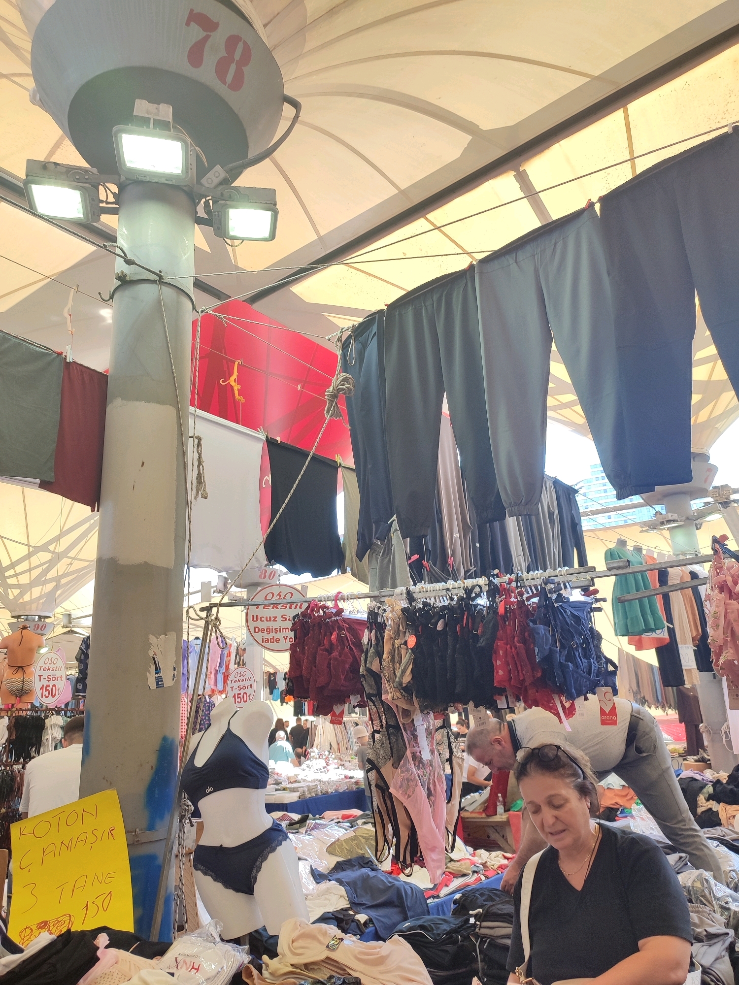 A busy outdoor market scene is depicted in Kadıköy, Turkey, with stalls displaying a variety of clothing items. In the foreground, a female shopper, wearing glasses and a black v-neck top, examines merchandise. Behind her, a man in a grey shirt and jeans is bent over, seemingly attending to his stall. The market is covered by a light-colored canopy. Clothing, including pants, underwear, and t-shirts, is hung from various racks and lines. A mannequin displays a dark blue bra and panty set. A yellow sign in Turkish is visible, with text including "KOTON ÇAMAŞIR" and prices. The overall impression is one of an active, commercial environment.