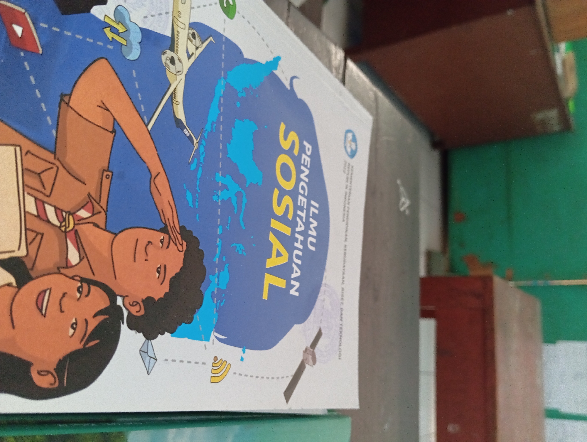 The image shows a close-up of the cover of a textbook titled "ILMU PENGETAHUAN SOSIAL." The cover features illustrations of two people, a satellite, an airplane, and a map of Indonesia. The book is placed on a table in what appears to be a classroom or school setting, as indicated by the presence of wooden cabinets and a green wall in the background. The lighting suggests it is daytime. The image does not provide any specific information or visual cues to determine the location within the city of Pandaan, Indonesia.