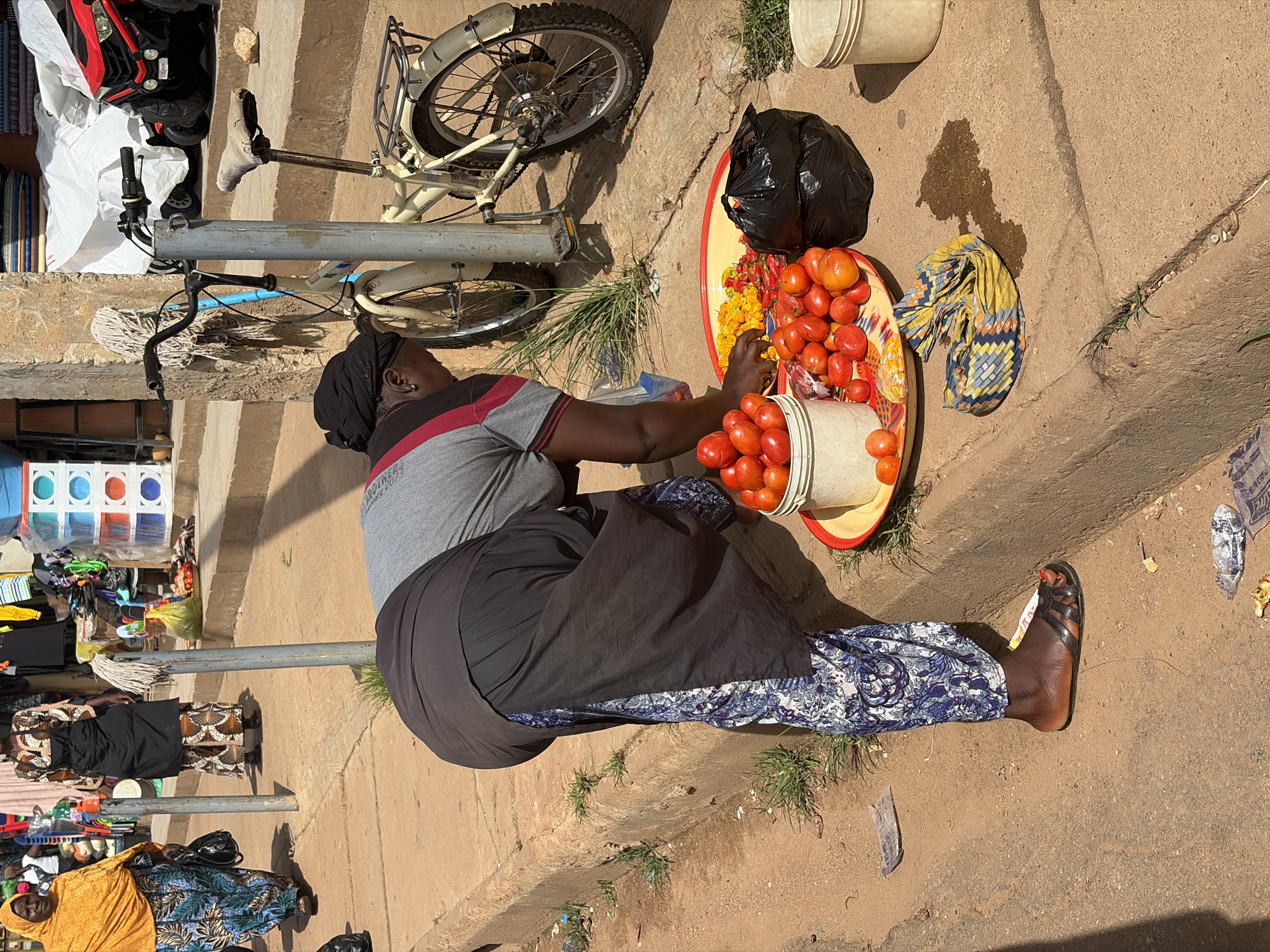 The image depicts a street market scene in Jos, Nigeria. A woman wearing a black headscarf and a grey and red striped shirt is bent over, arranging produce. She is wearing a dark-colored skirt and a patterned blue and white fabric wraps around her legs. In front of her, on a red and yellow tray, are piles of tomatoes and what appear to be chili peppers or other small, colorful vegetables. A white bucket filled with tomatoes is to the left of the tray, and a black plastic bag is positioned to the right. Another white bucket is visible further to the right. A bicycle with a mop tied to its handlebars stands behind the woman. In the background, other market stalls and people are visible, including a woman in an orange headscarf and a patterned dress walking away from the camera. The ground is a mixture of dirt and concrete, and patches of grass grow along the curb.
