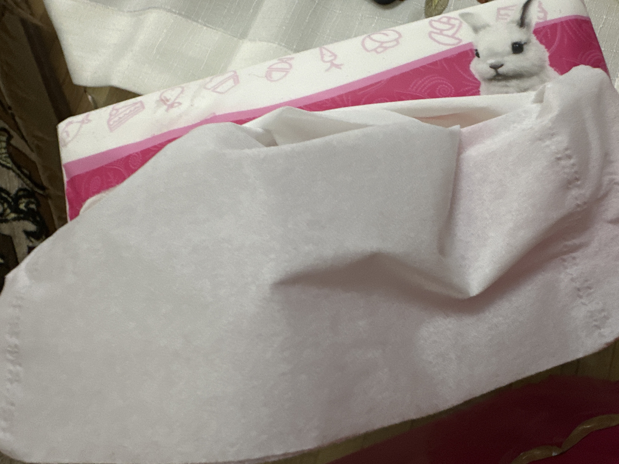 A close-up, angled shot shows a tissue box peeking out from under a white tissue. The tissue box has a pink and white striped design with small cartoon images of cupcakes, strawberries, and what appear to be rabbits. One rabbit's face is visible, looking out from under the tissue. The background is blurred and appears to be a patterned surface, possibly upholstery. The image does not provide any information about its location in Yerevan, Armenia.