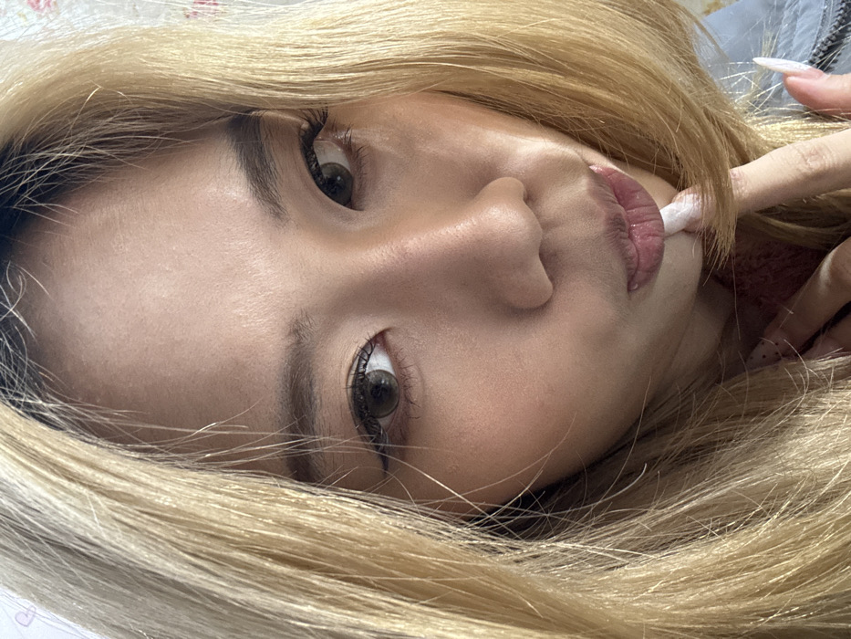 The image displays a close-up, head-and-shoulders portrait of an individual, identified by the prompt as being in Shapingba District, China. The subject has fair skin and long, blonde hair with visible darker roots. The individual looks directly at the camera with a subtle expression. Their eyes feature dark brown irises, possibly enhanced by colored contact lenses, and are lined with dark eyeliner and prominent eyelashes. Eyebrows are dark and well-defined. The nose is centrally prominent. The lips are full and glossy, with the tip of a long, white, stiletto-shaped fingernail from the individual's right hand gently touching the lower lip area. Other long, white fingernails are partially visible on the same hand. The subject wears a reddish garment visible at the neck, and a light blue/grey garment is visible on the right side of the frame. The background is light-colored, suggesting an indoor setting, with a faint floral pattern visible on the upper right side.