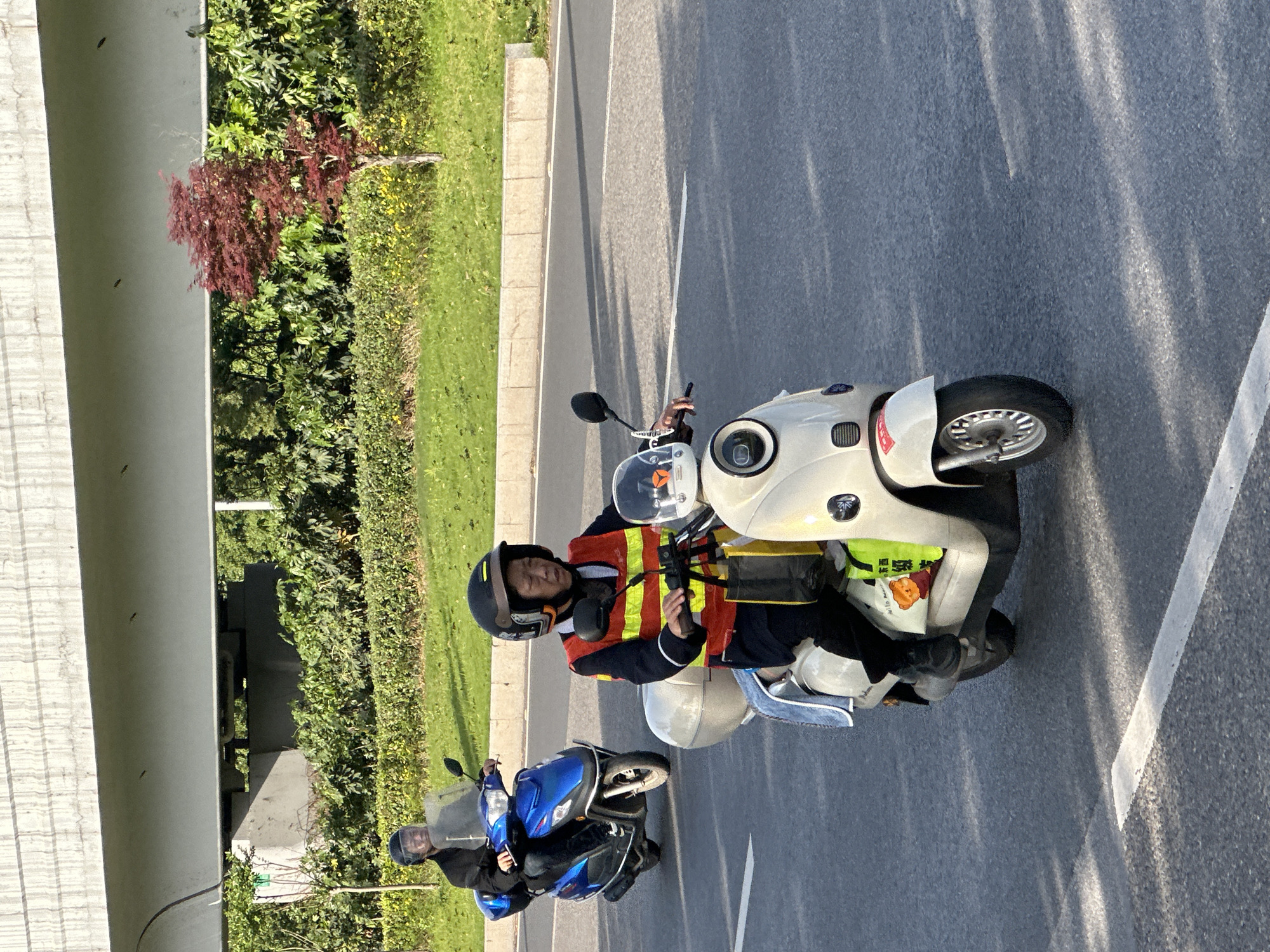 The image displays two individuals on scooters on a paved road in what appears to be an urban or suburban environment in Jiangjin, China, under clear, sunny daytime conditions with long shadows indicating an angled sun.
In the foreground, a person, likely male, is seated on a light-colored (off-white/cream) electric scooter. They are wearing a dark helmet, a dark long-sleeved top, and a bright orange and yellow reflective vest. This individual is looking towards the left, holding an object, possibly a mobile phone or radio, in their right hand. A yellow bag, featuring a cartoon bear and partial Chinese text that includes "My Life..." and "欣怡" (Xīnyí), hangs from the scooter's handlebars.
Slightly behind and to the left, another person, also wearing a dark helmet and dark clothing, is riding a blue electric scooter. This person is looking forward. Both scooters are equipped with windshields.
The road features white painted lines, suggesting a designated lane or shoulder. Along the right side of the road (from the viewer's perspective), there is a lush green verge with grass, various shrubs, and trees, including one with distinctive dark red foliage. A large, grey concrete structure, resembling an overpass or bridge, is prominent on the left side of the frame, hinting at multi-level infrastructure. The scene depicts everyday transit or a paused journey in a public area.